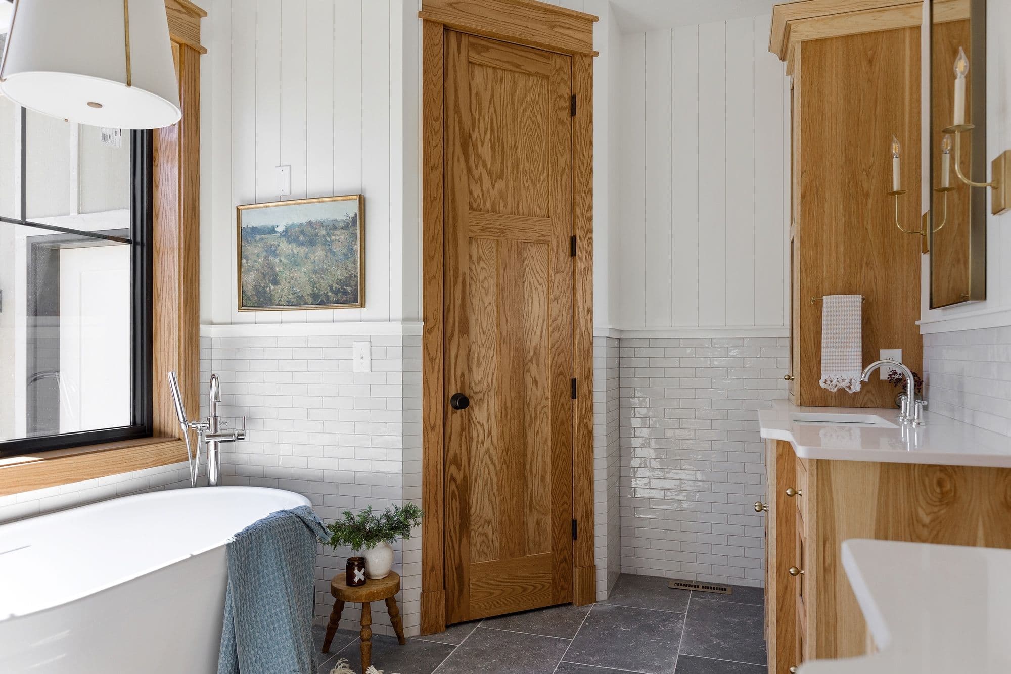 Bathroom featuring white subway tile, wood vanity, and a freestanding tub under a black-framed window. The flooring is dark gray stone, and there's a warm wood door with matching trim.