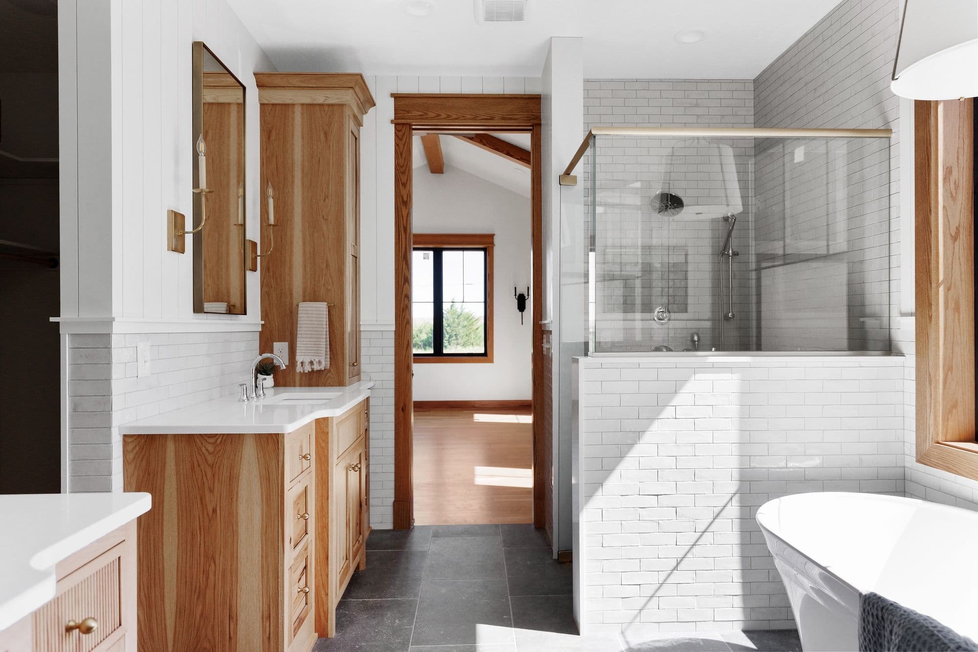 Bathroom featuring a wood vanity with white countertop, wood-framed mirror, a glass-enclosed shower with white subway tile, and a freestanding bathtub. The floor is dark gray tile, and a wood-framed doorway leads to another room.