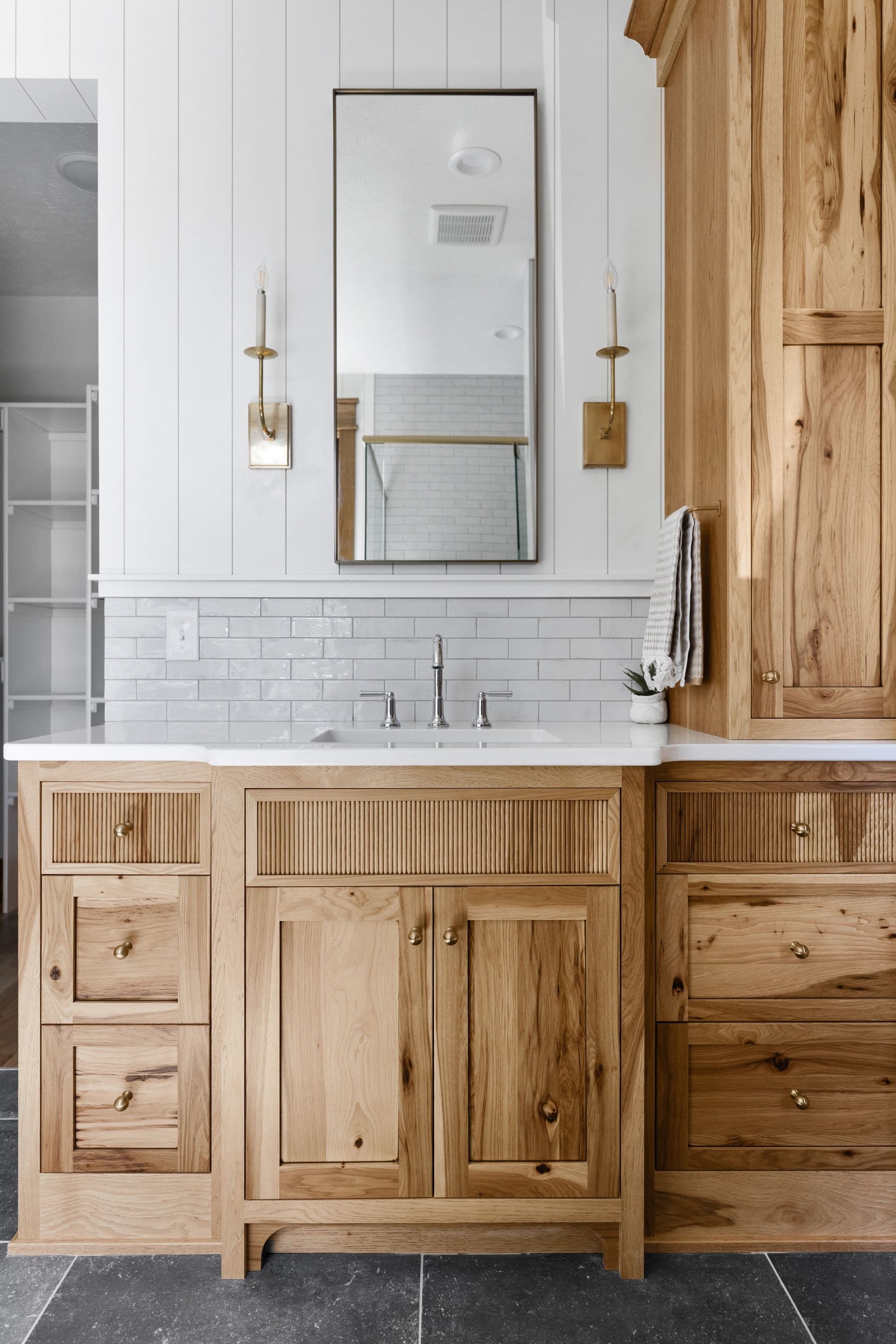 Bathroom vanity featuring light wood cabinetry with reeded drawer fronts and brass hardware. A white countertop and subway tile backsplash complement the shiplap walls and brass sconces.