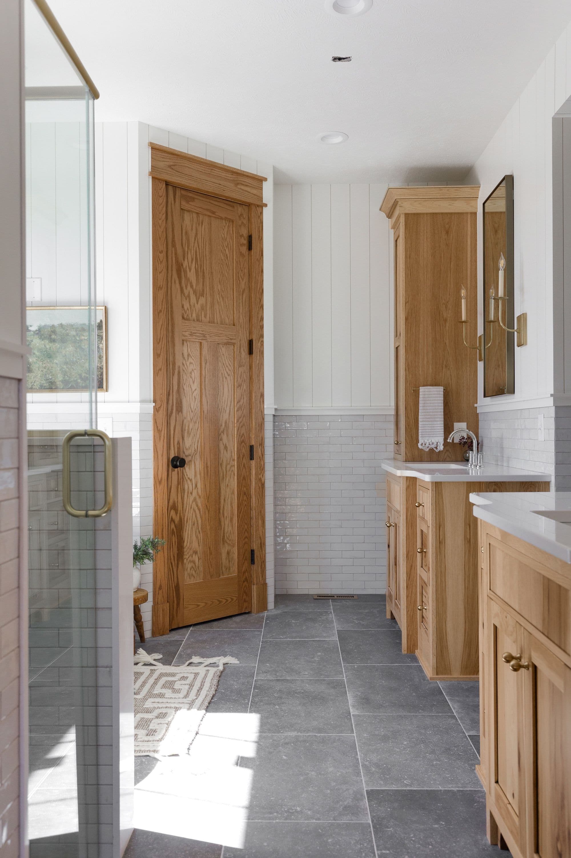 Bright bathroom with light wood cabinetry, gray tile flooring, and white shiplap walls. A partially open wooden door reveals a small rug on the floor, with a glass-enclosed shower to the left.