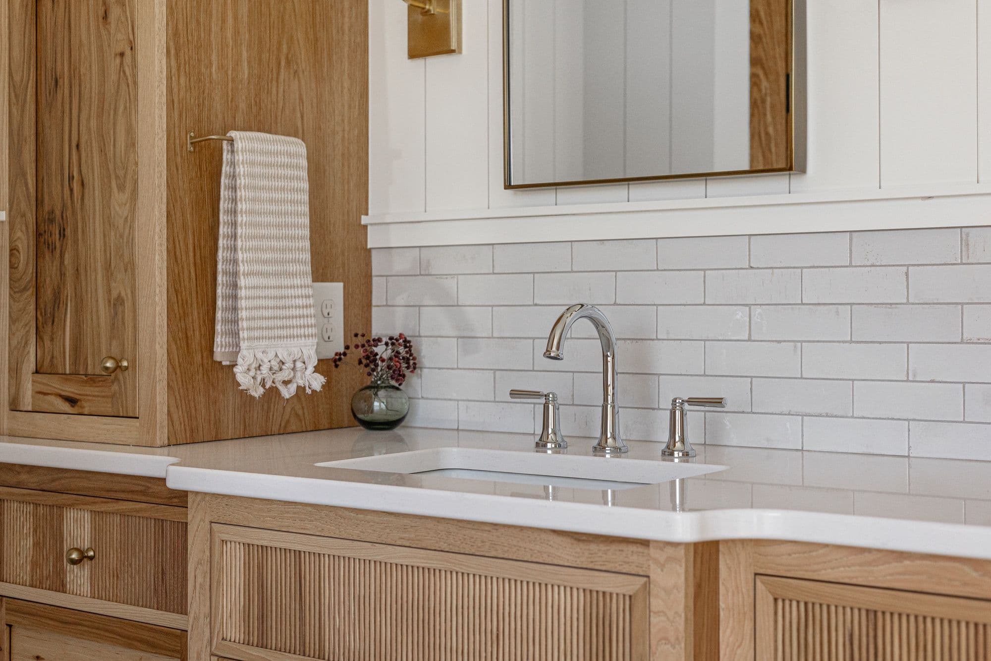 Bathroom vanity features light wood cabinets with reeded fronts, a white countertop with an undermount sink, and chrome fixtures. A gray and white towel hangs on a gold rod.