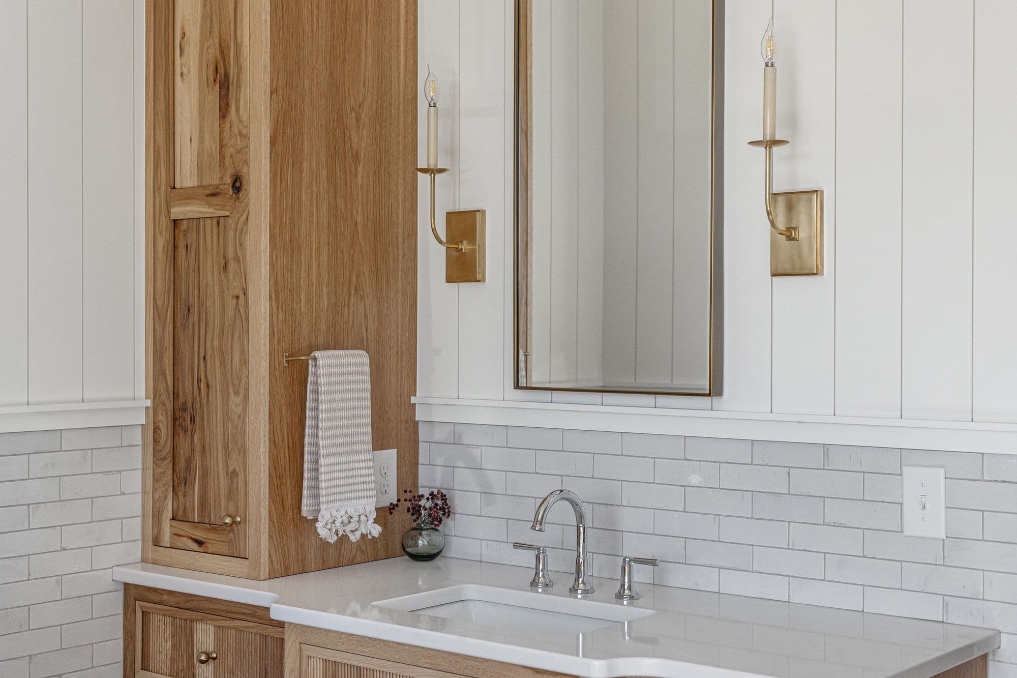 Bathroom vanity with a white countertop, a rectangular undermount sink, and polished chrome faucet. A light wood cabinet sits on the counter to the left of the mirror, which is lit by wall sconces with candlestick lights.