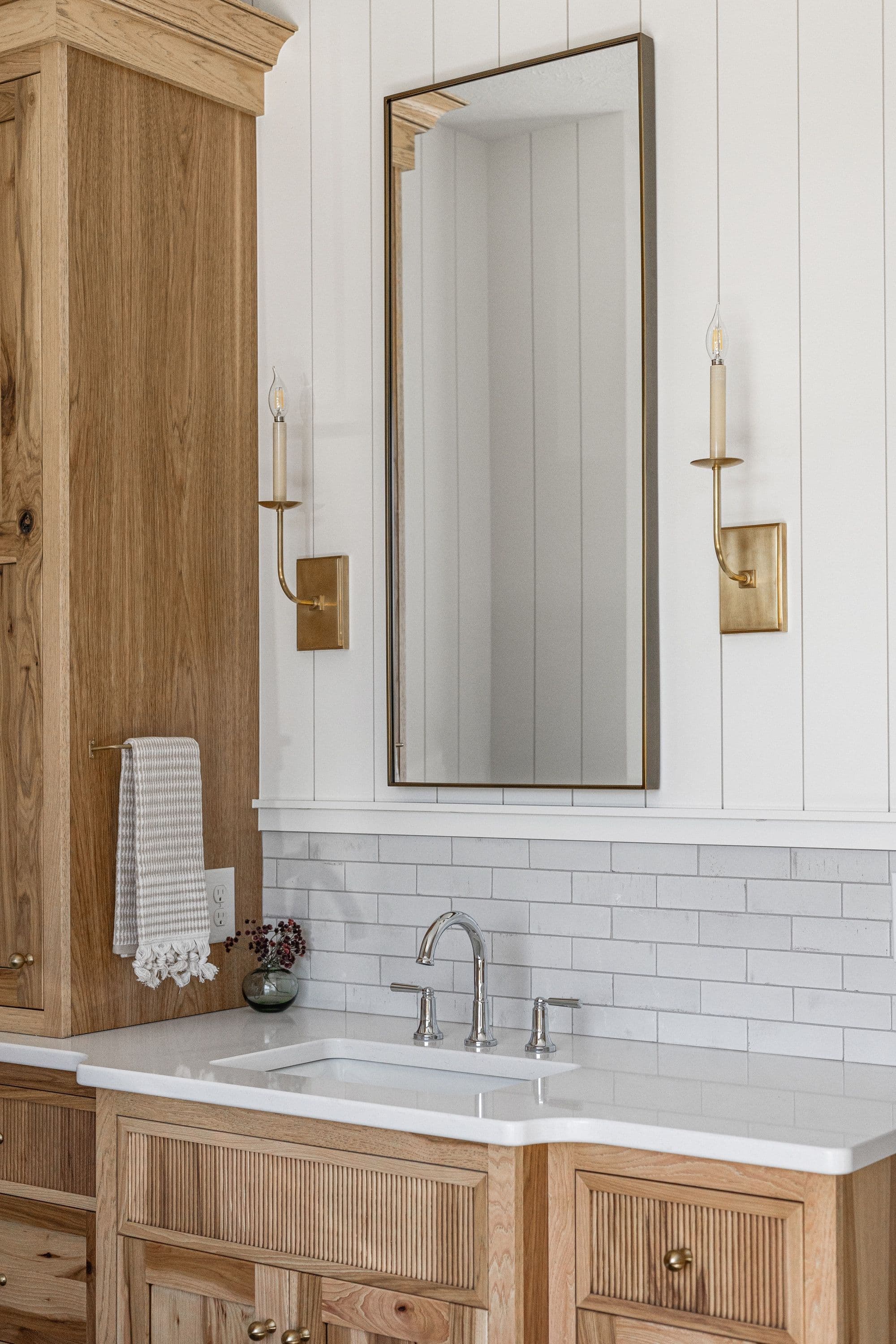 Bathroom features a wood vanity with fluted detailing, white countertop, and subway tile backsplash. A rectangular mirror hangs above the sink, flanked by sconce lighting and white vertical paneling.