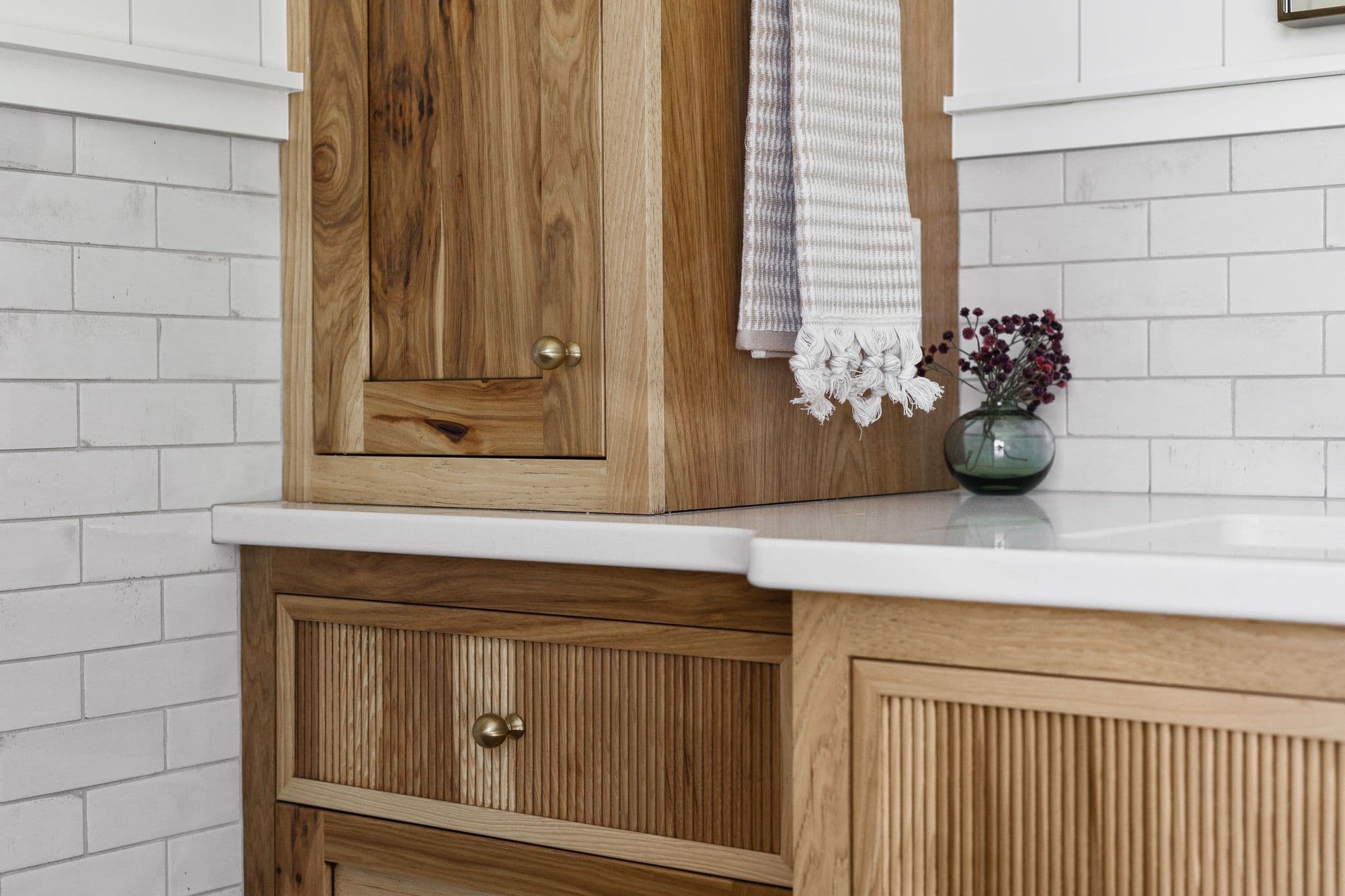 Close-up of a bathroom vanity with natural wood cabinets and drawers featuring vertical reeding. White quartz countertops and a subway tile backsplash create a clean, modern aesthetic.