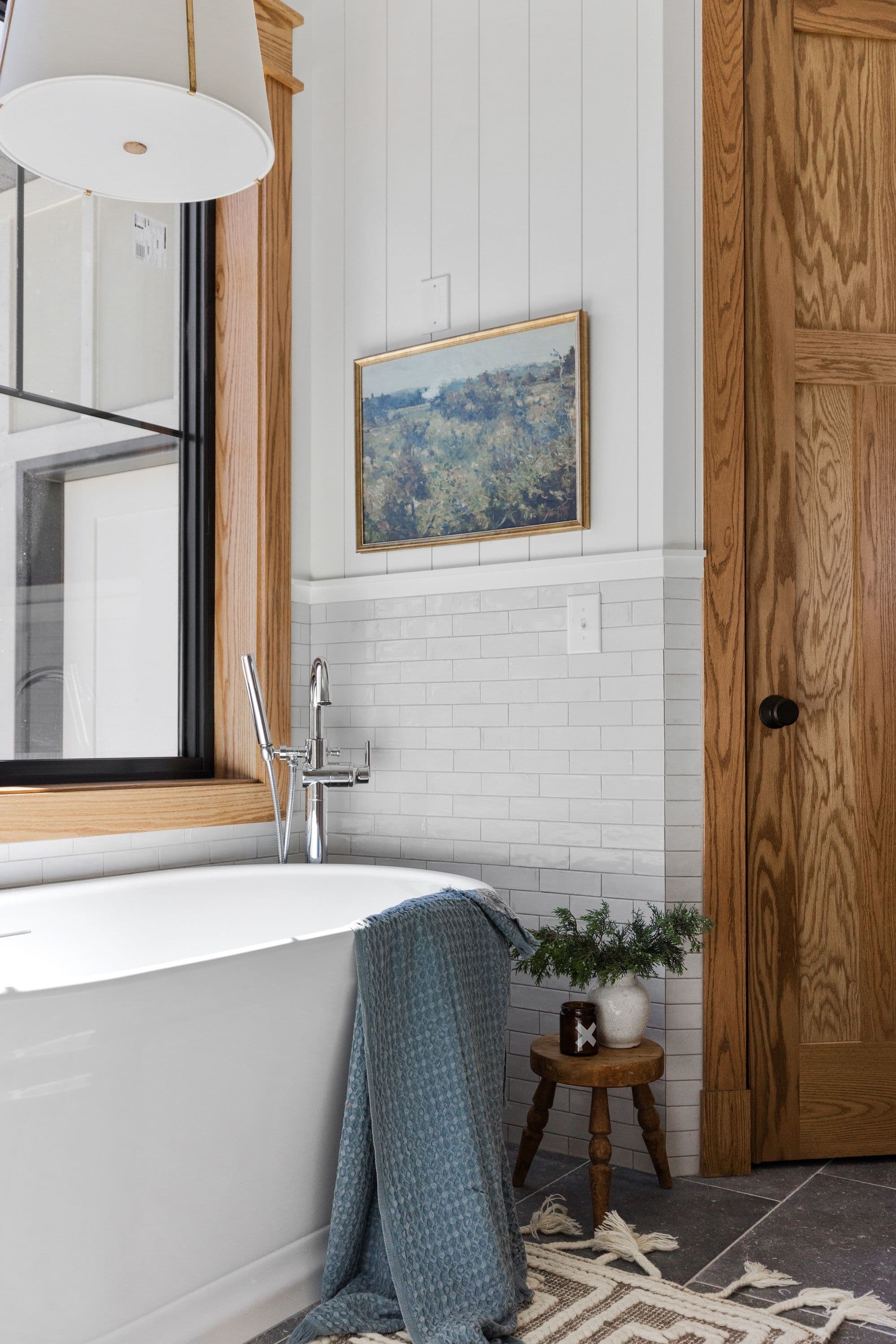Bathroom featuring a white freestanding tub with chrome faucet and shower wand, light wood-framed window and door, white subway tile and vertical plank walls, and a framed landscape painting. A small wooden stool with a white vase and green sprigs sits beside the tub.