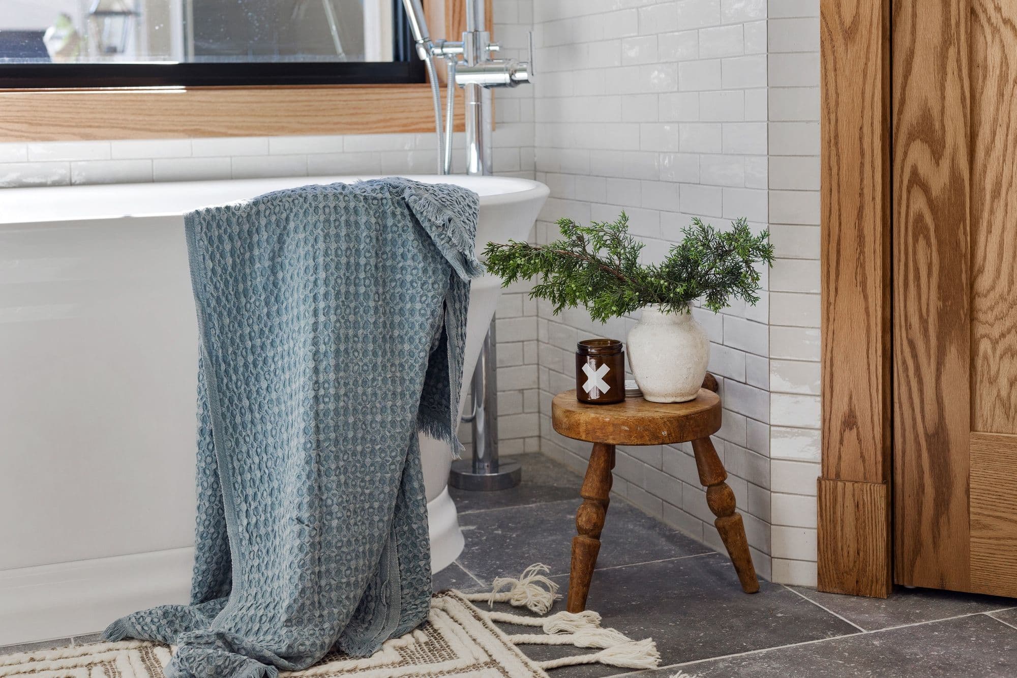 Bathroom with white subway tile walls and a freestanding tub is adorned with a blue textured towel. A rustic wooden stool next to the tub holds a white vase with greenery and a candle.