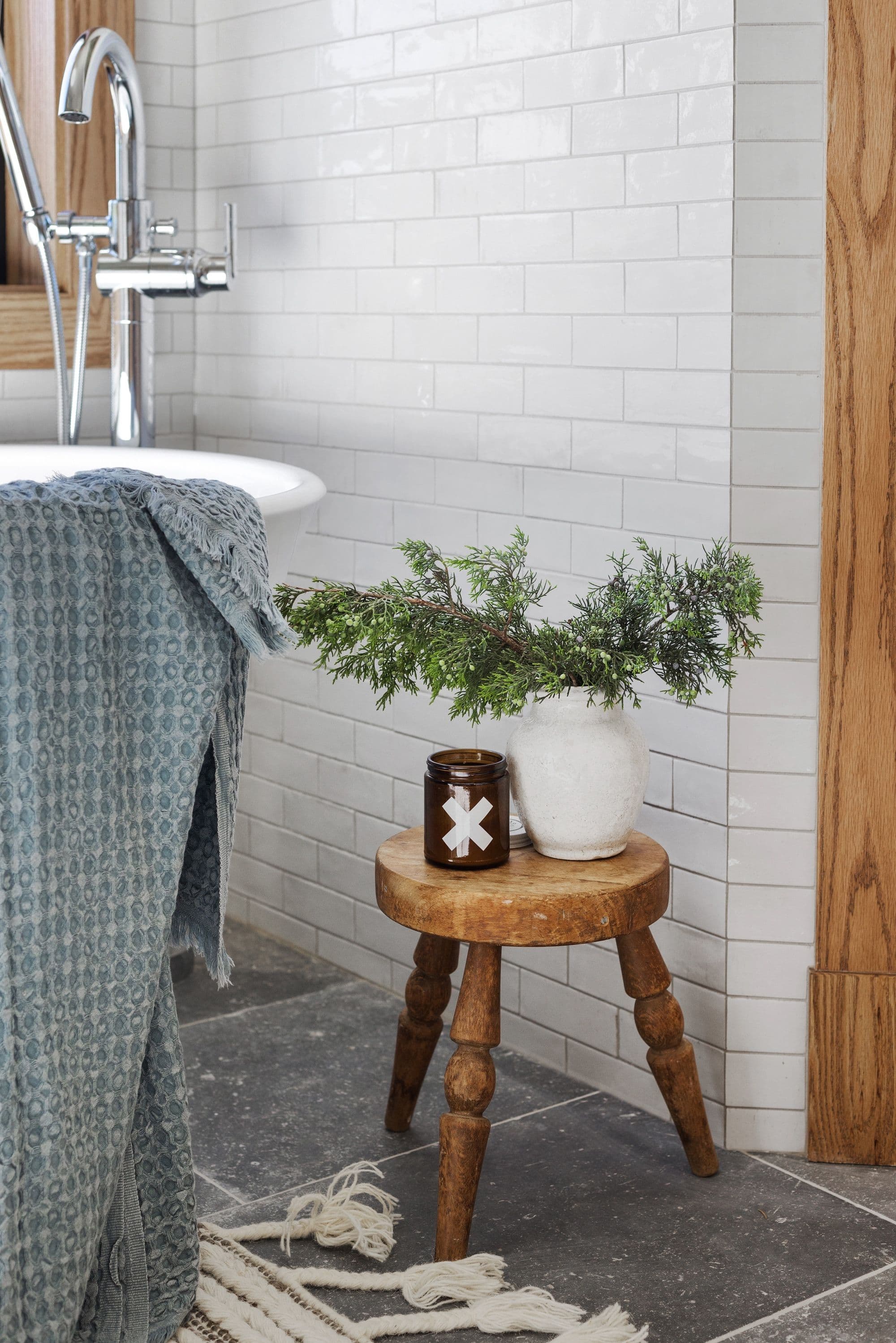 Bathroom features white subway tile walls, a freestanding tub with a chrome faucet, and a wooden stool holding a vase with greenery and a candle. A blue waffle-weave towel drapes over the side of the tub.
