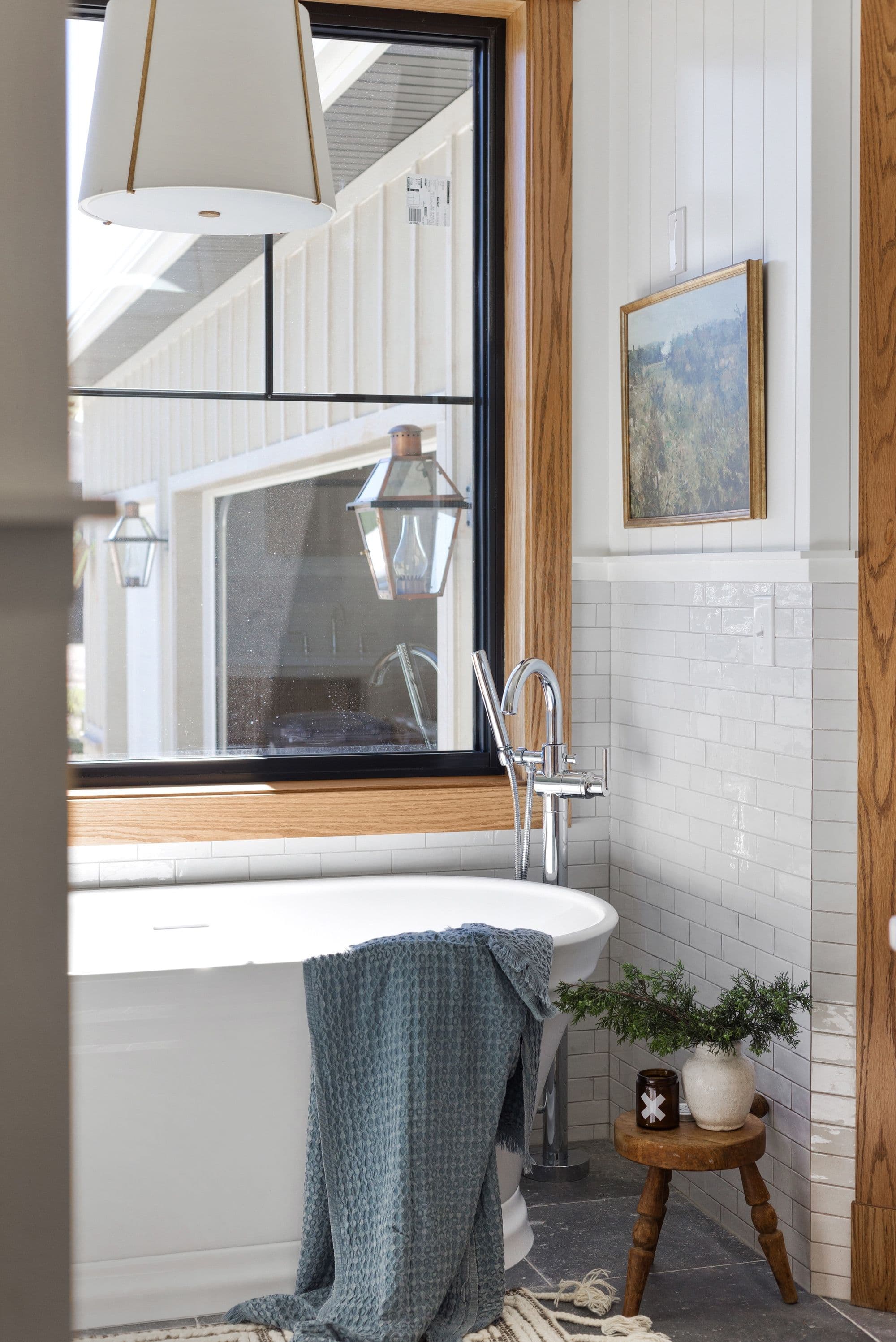 Bathroom features a soaking tub with a blue waffle-weave towel draped over its side, complemented by a wooden stool holding greenery and a small jar, set against a backdrop of white subway tile and board and batten walls. A black-framed window offers a glimpse of the exterior, reflecting hanging lanterns.