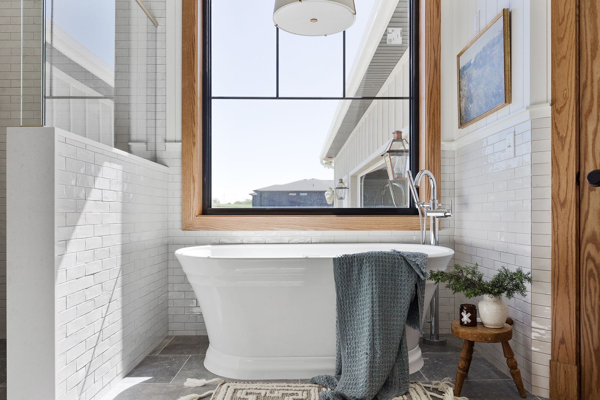 Bright bathroom featuring a freestanding white tub with a blue towel draped over it, set against a backdrop of white subway tile walls. A wooden-framed window provides natural light, and a small wooden stool holds a vase with greenery and a candle.