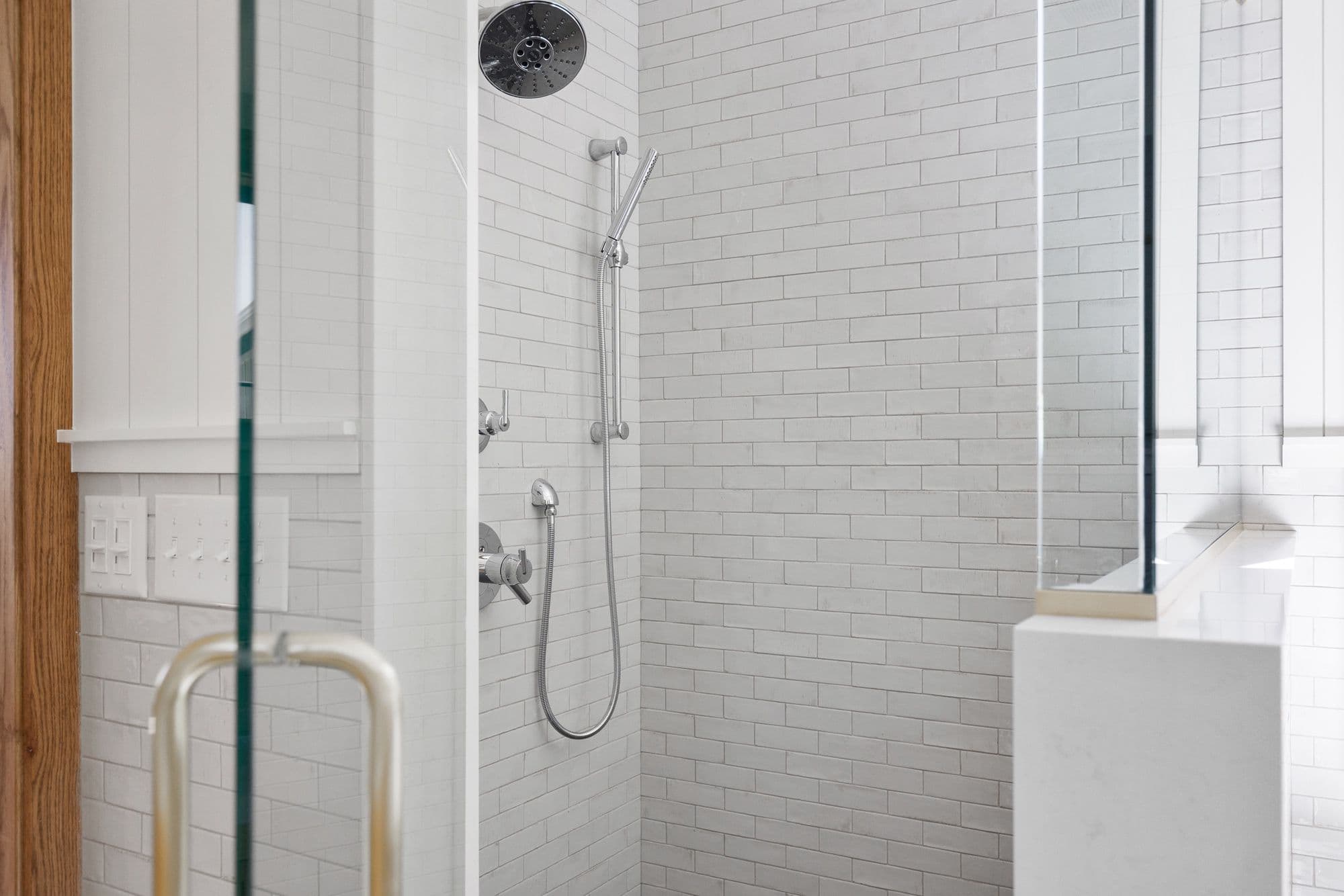 Modern bathroom features a glass-enclosed shower with white subway tile and chrome fixtures. There is a wood door frame adjacent to the shower with a white wall containing light switches outside the shower.