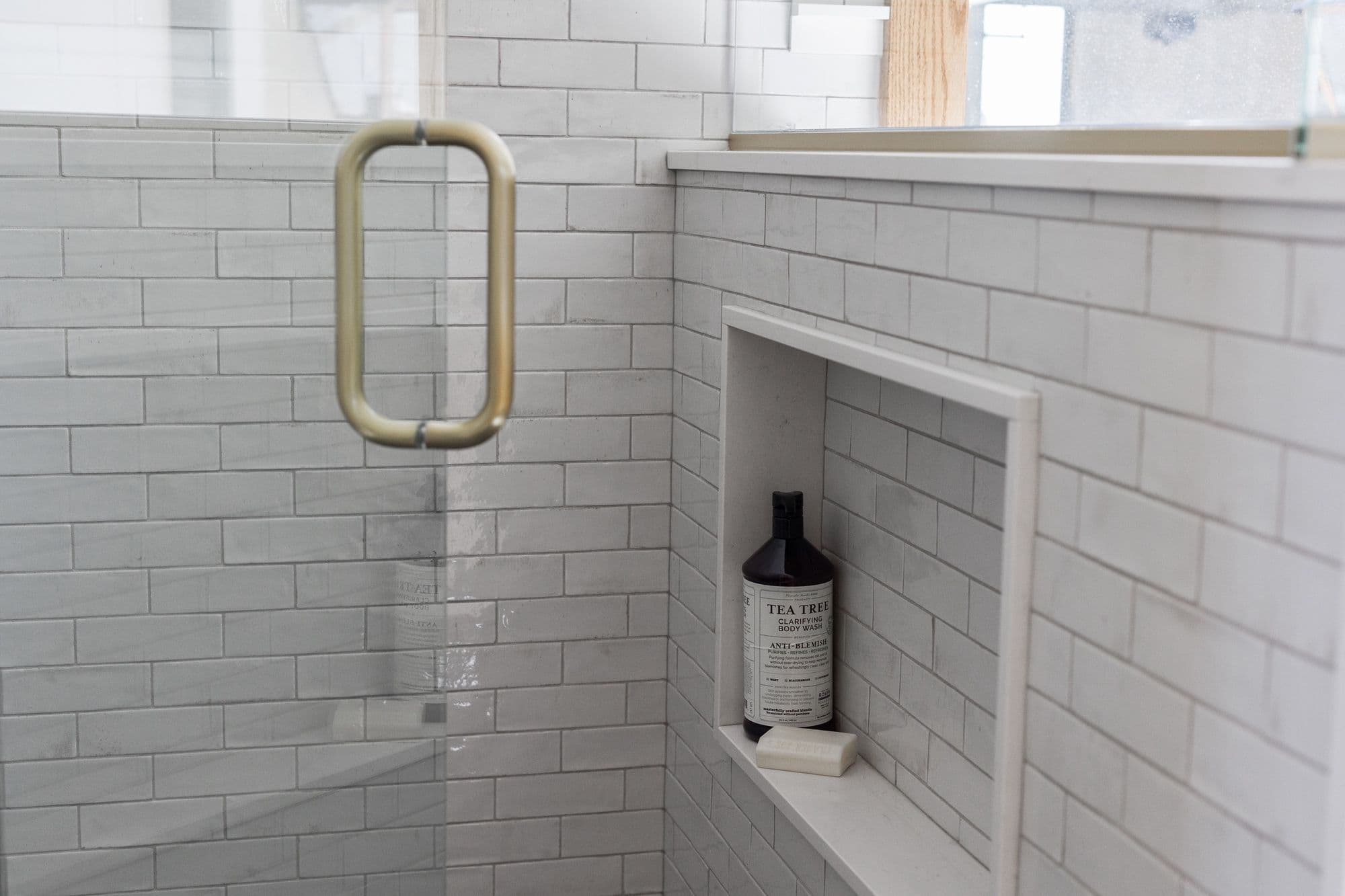 Shower with glass door and brass handle features a built-in niche with white subway tile and a bottle of tea tree body wash. The bathroom's clean aesthetic is highlighted by the light gray grout and soft lighting.