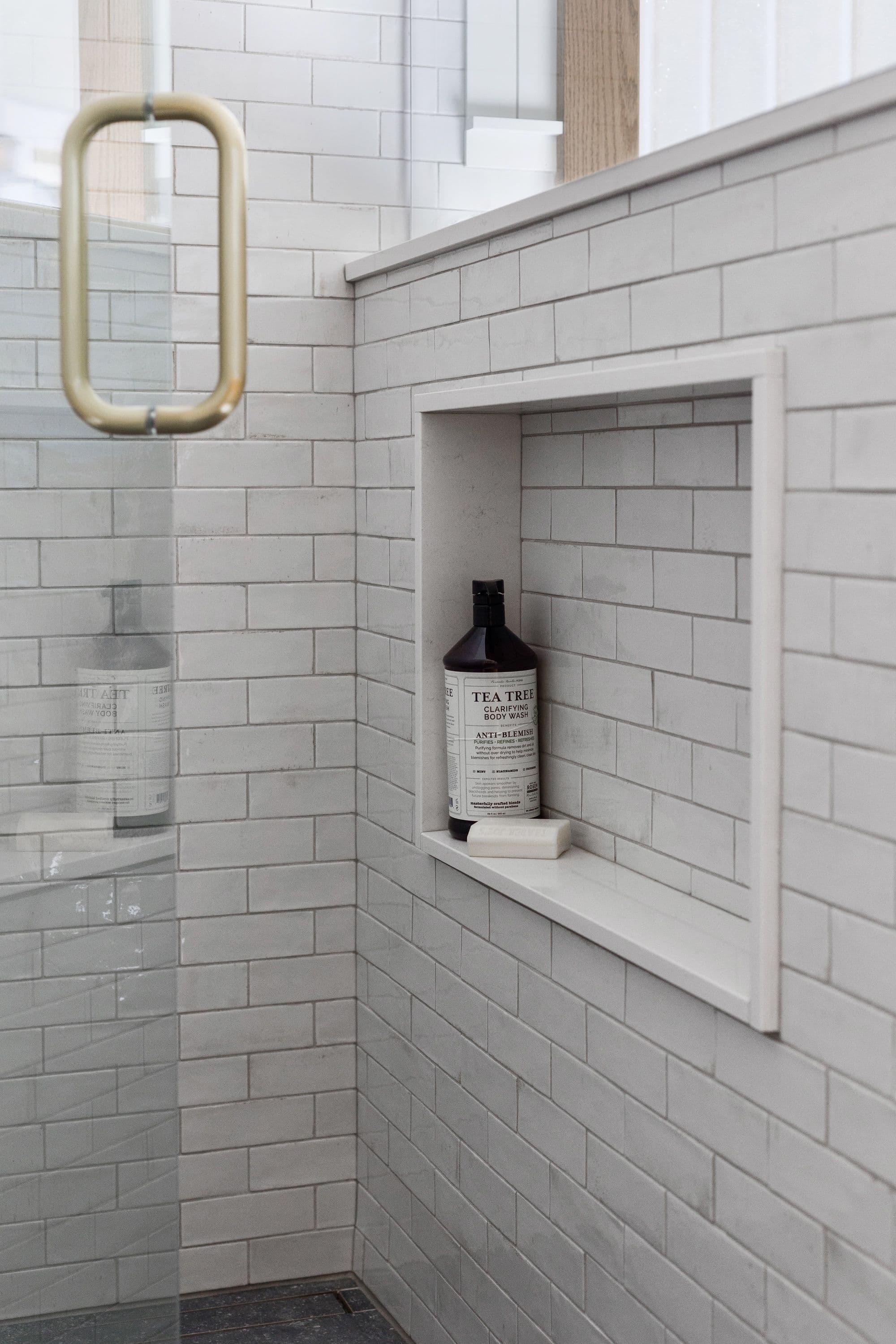 This bathroom features a shower area with a built-in niche for storage, filled with a bottle of tea tree body wash and a bar of soap. The walls are covered in light gray subway tiles, and the shower has a glass door with a gold handle.