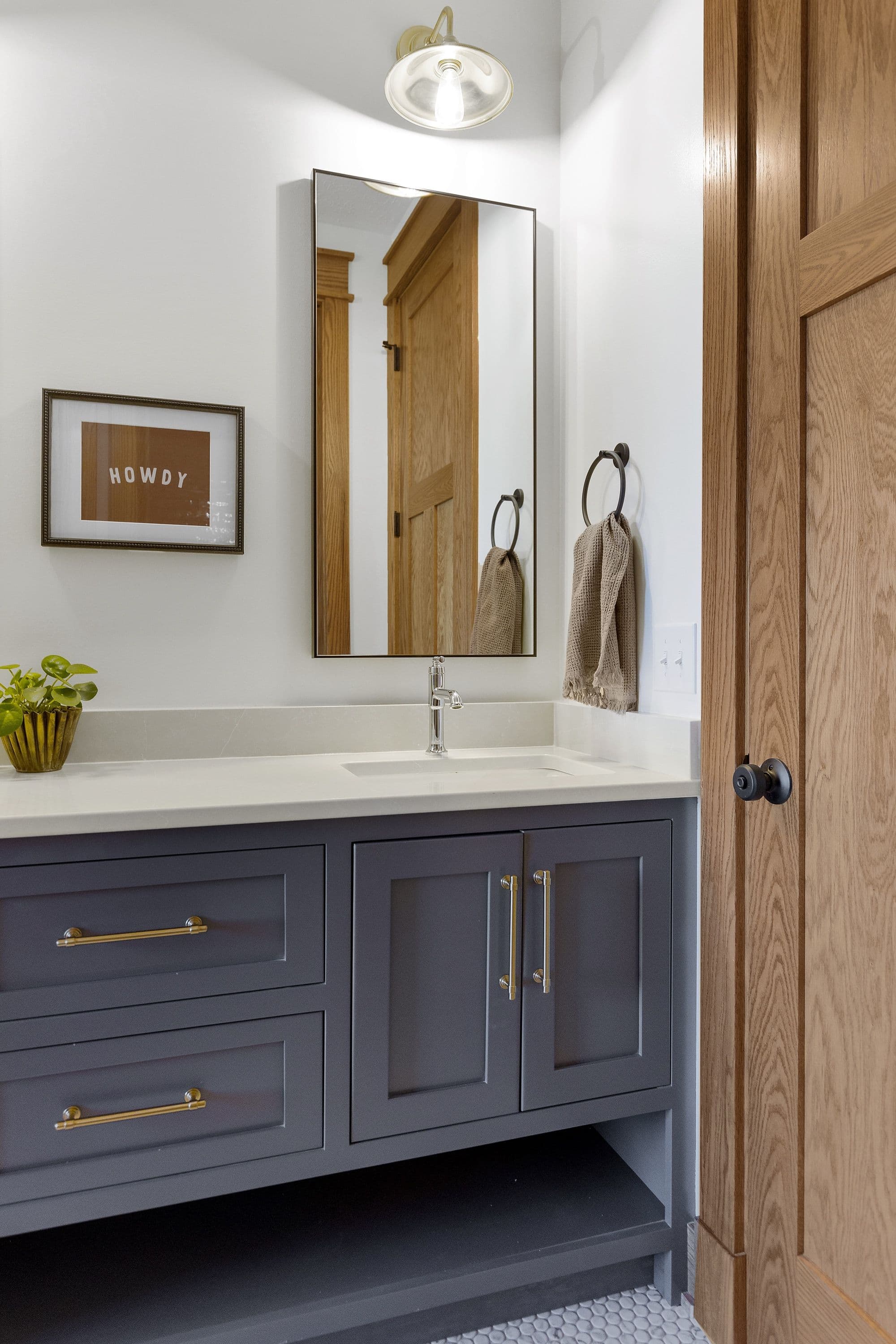 Bathroom features a gray vanity cabinet with gold hardware, topped with a light countertop and undermount sink. A black-framed mirror hangs above, reflecting a wooden door, and a framed "HOWDY" print adds a rustic touch to the white walls.