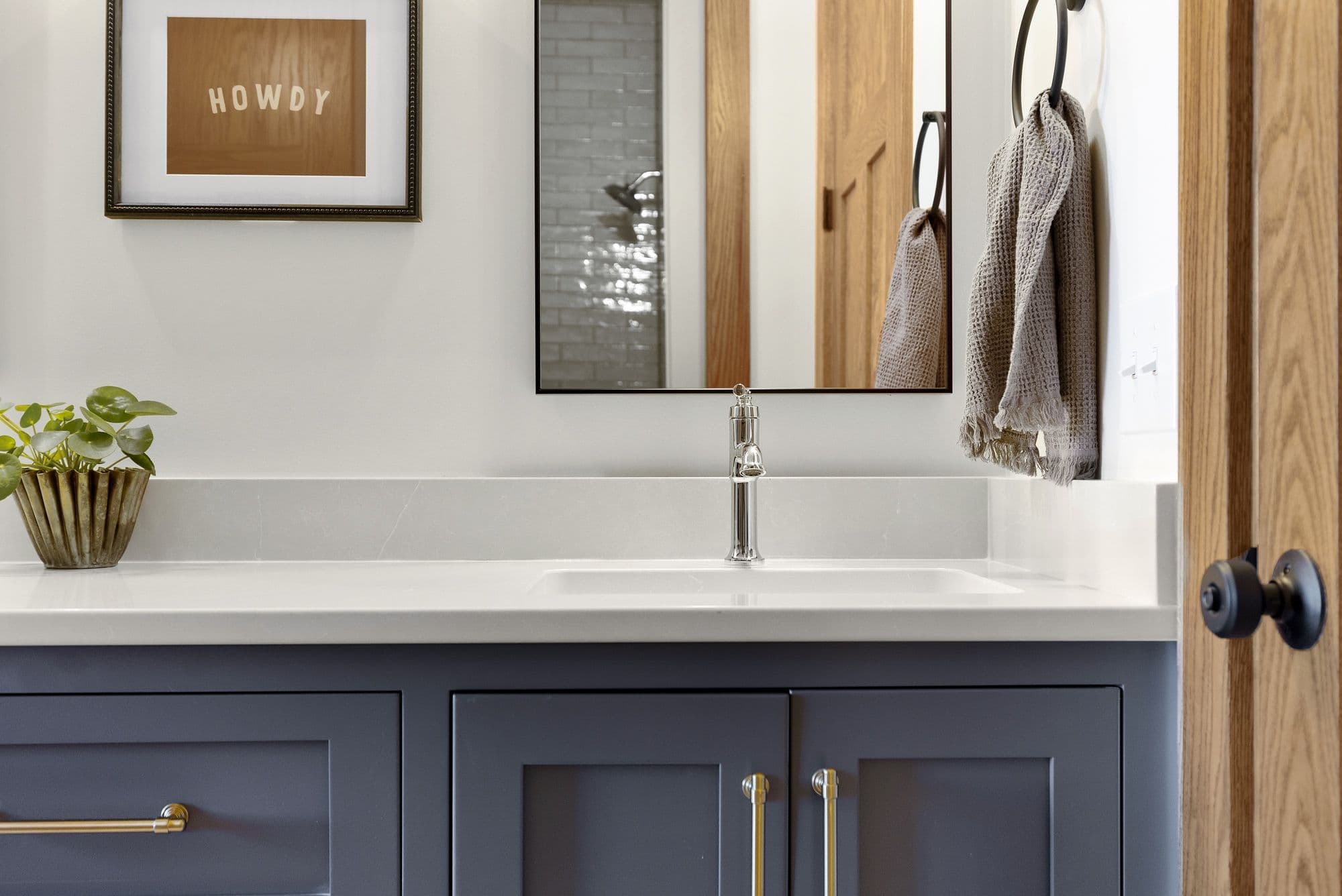 Bathroom features a gray vanity with white countertop, chrome faucet, and rectangular mirror. A framed art piece with the word 'HOWDY' hangs above a decorative plant, with a wood door and towel ring visible to the side.