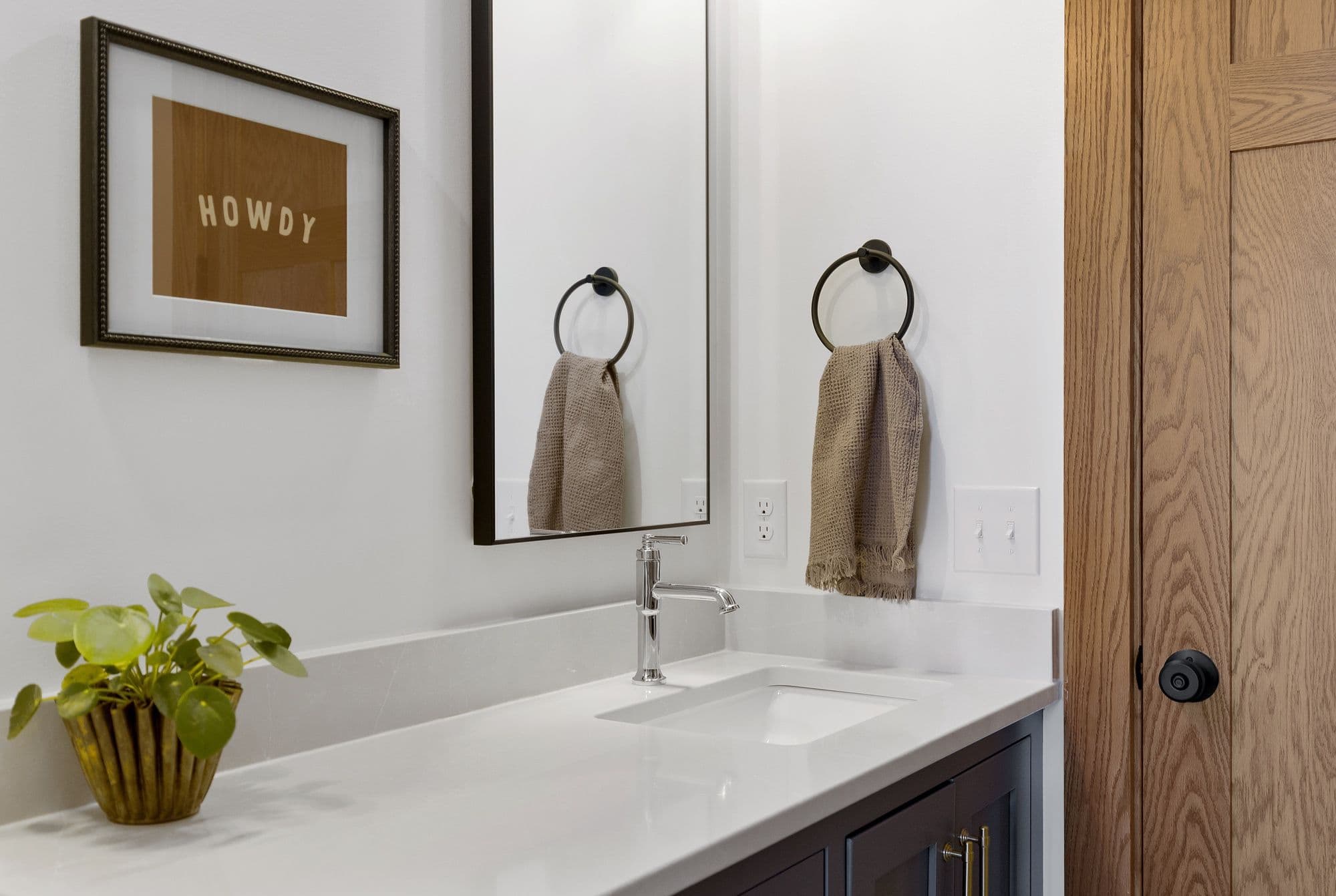 Bathroom interior featuring a white countertop with integrated sink, chrome faucet, black framed mirror, and a framed "HOWDY" print on the wall. The design includes a dark vanity cabinet with gold hardware, light wood door, and a potted plant.