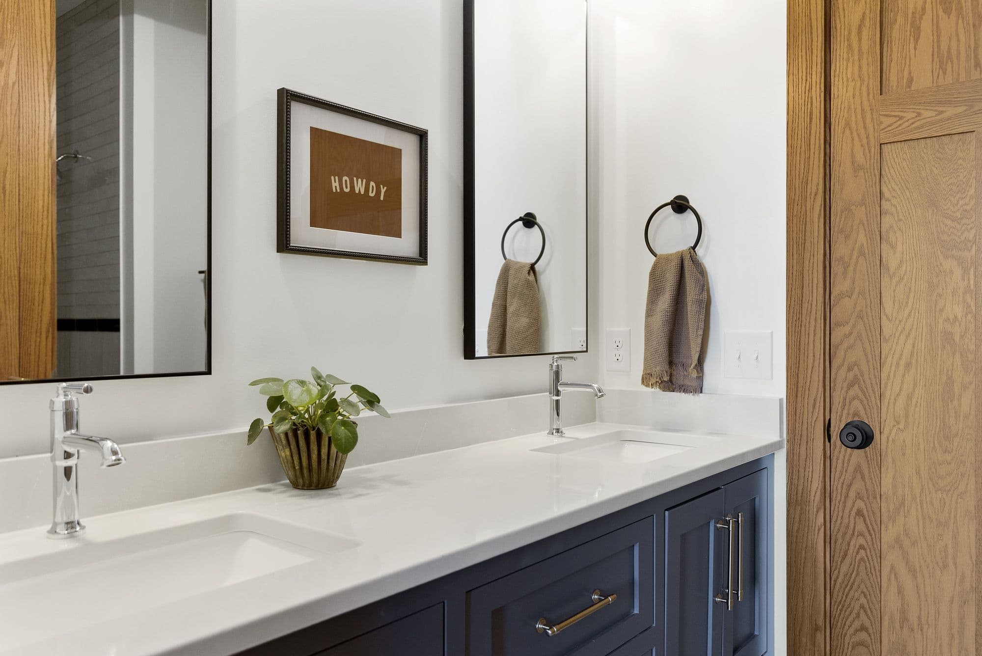 Bathroom featuring a double vanity with white countertops and navy blue cabinets, flanked by mirrors and artwork. The space includes chrome fixtures, towel rings, and a potted plant.