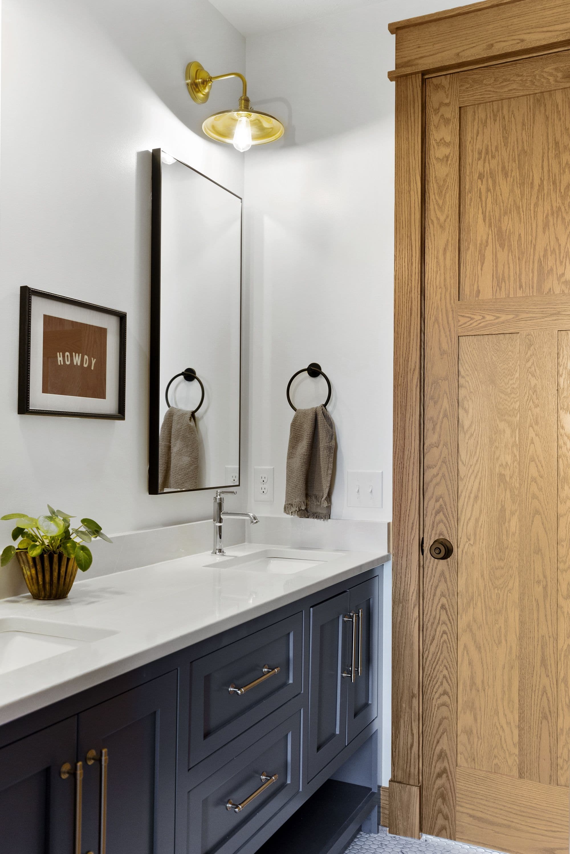 Bathroom interior features a vanity with white countertop and gray cabinetry with brass hardware. A mirror hangs above the sink, flanked by a 'Howdy' sign and a brass sconce lighting fixture.
