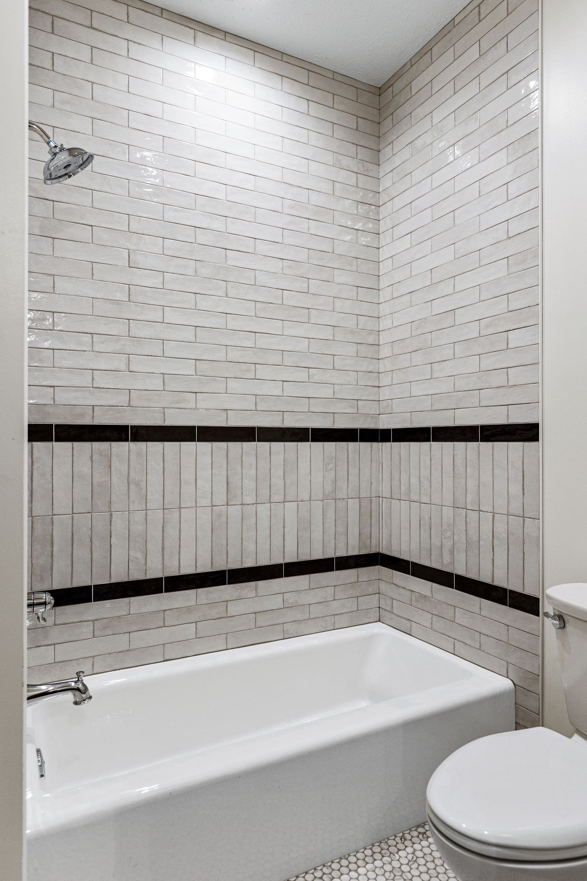 Bathroom features a white bathtub with chrome fixtures, surrounded by textured gray and white subway tile walls with two black accent stripes. A white toilet is visible adjacent to the tub.