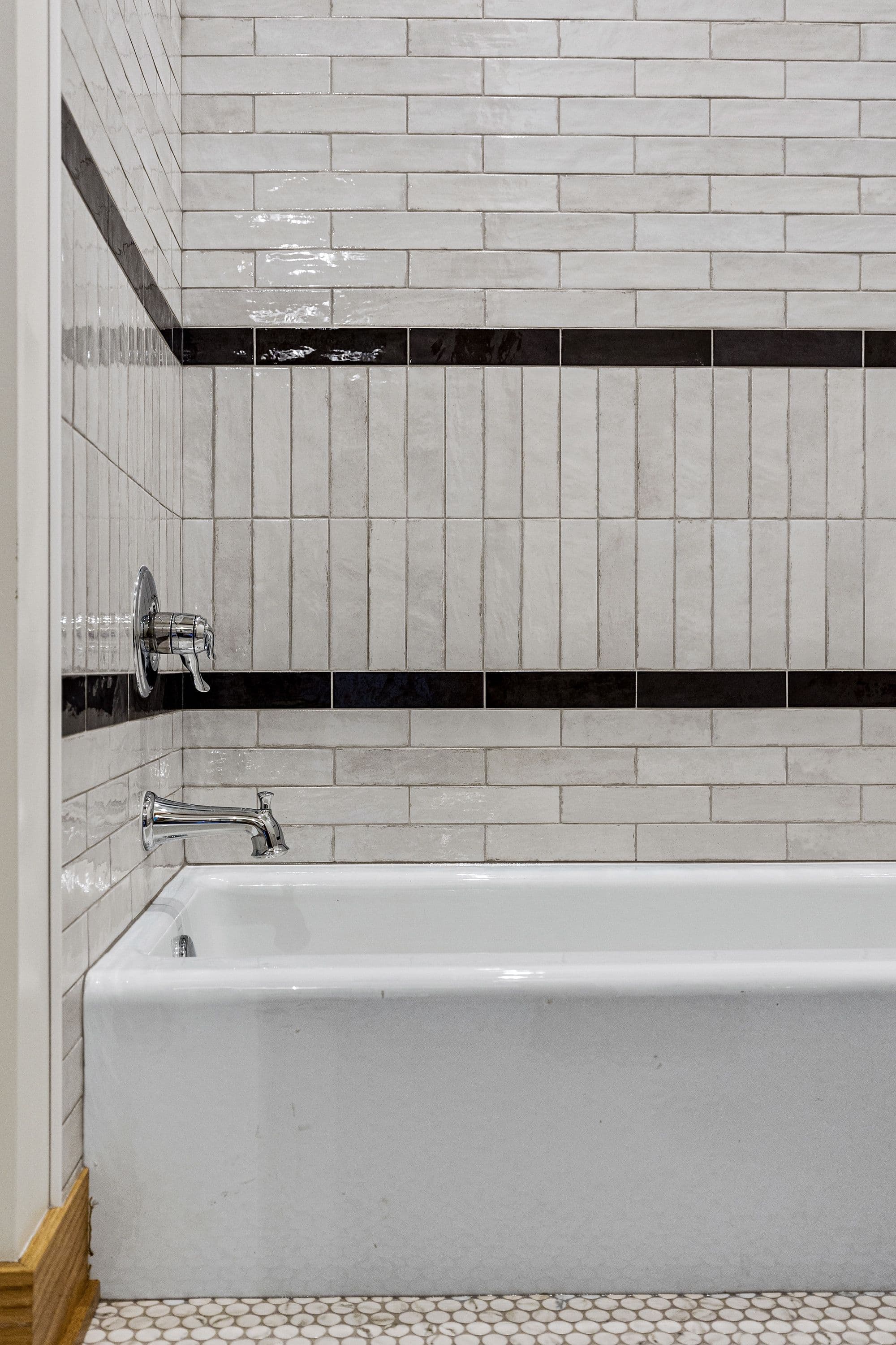 Bathroom featuring a white bathtub with chrome fixtures and a white tile wall accented with black tile stripes. The floor is tiled with white and gray mosaic tiles.