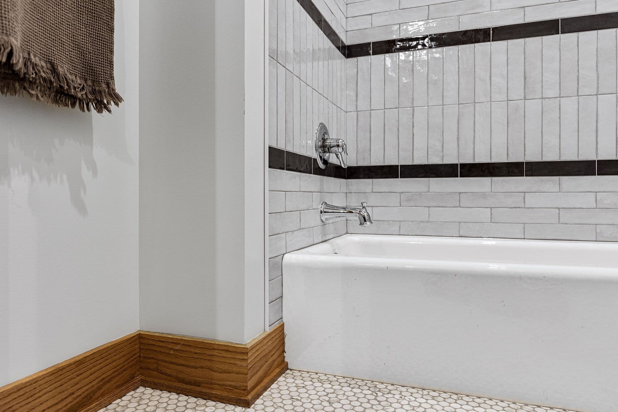 This bathroom features a white bathtub with chrome fixtures and light gray subway tile with a black horizontal stripe. The flooring consists of small, round, white tiles and a stained wood baseboard.