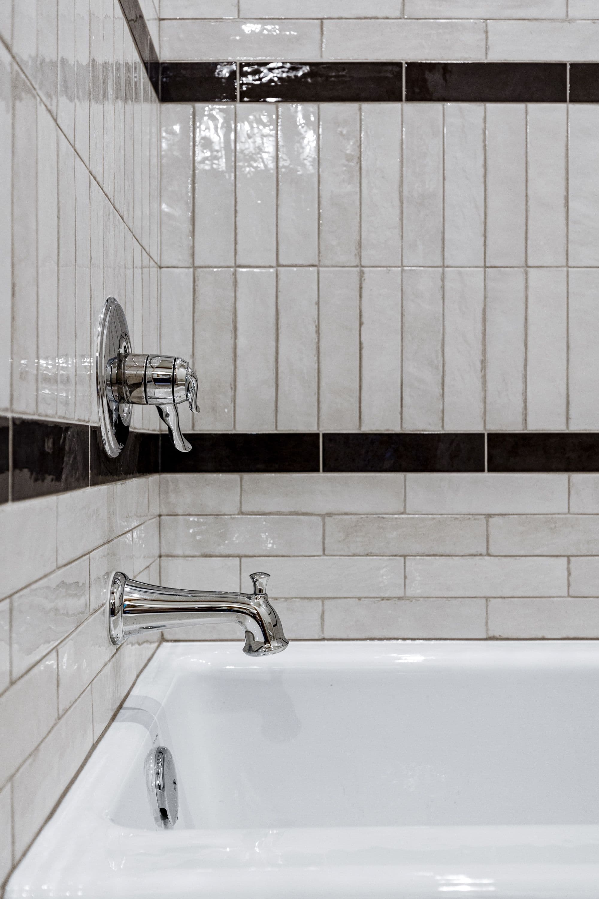This is a close up shot of a bathroom featuring a white tub and chrome fixtures. The walls are covered in light, textured tiles with a contrasting dark band running horizontally.