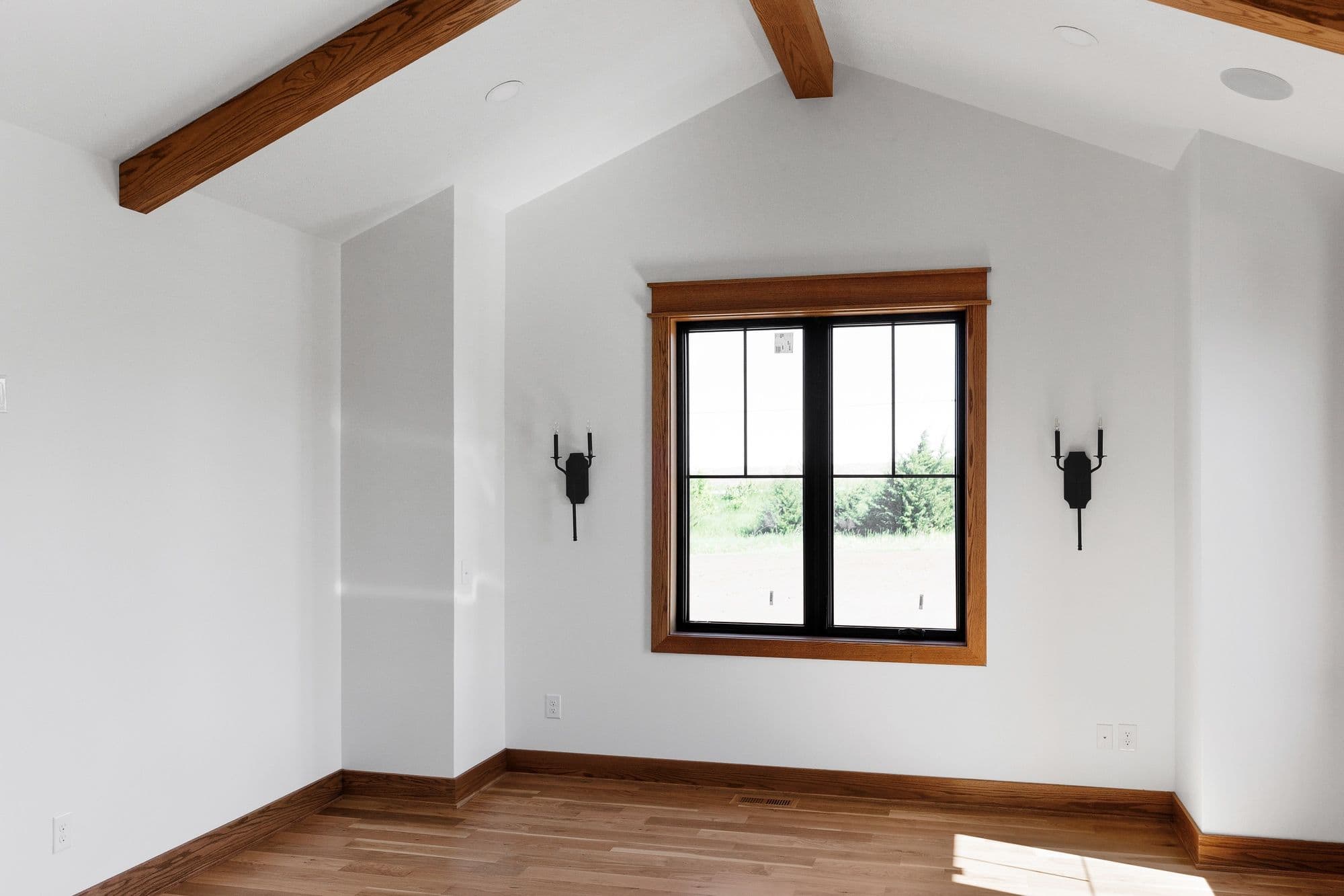 Bright room with hardwood floors and a black-framed window, featuring white walls and natural wood trim, as well as wooden ceiling beams. Black sconces flank the window, adding to the room's modern yet rustic aesthetic.