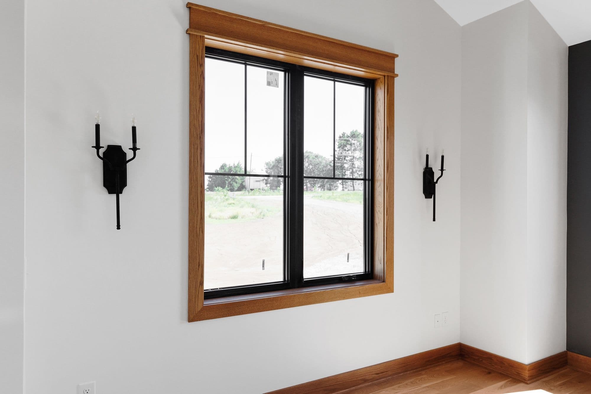 Interior shot of a room featuring a window with wooden trim and black framing that looks out onto a rural landscape. Two black sconces flank the window, mounted on a soft white wall.