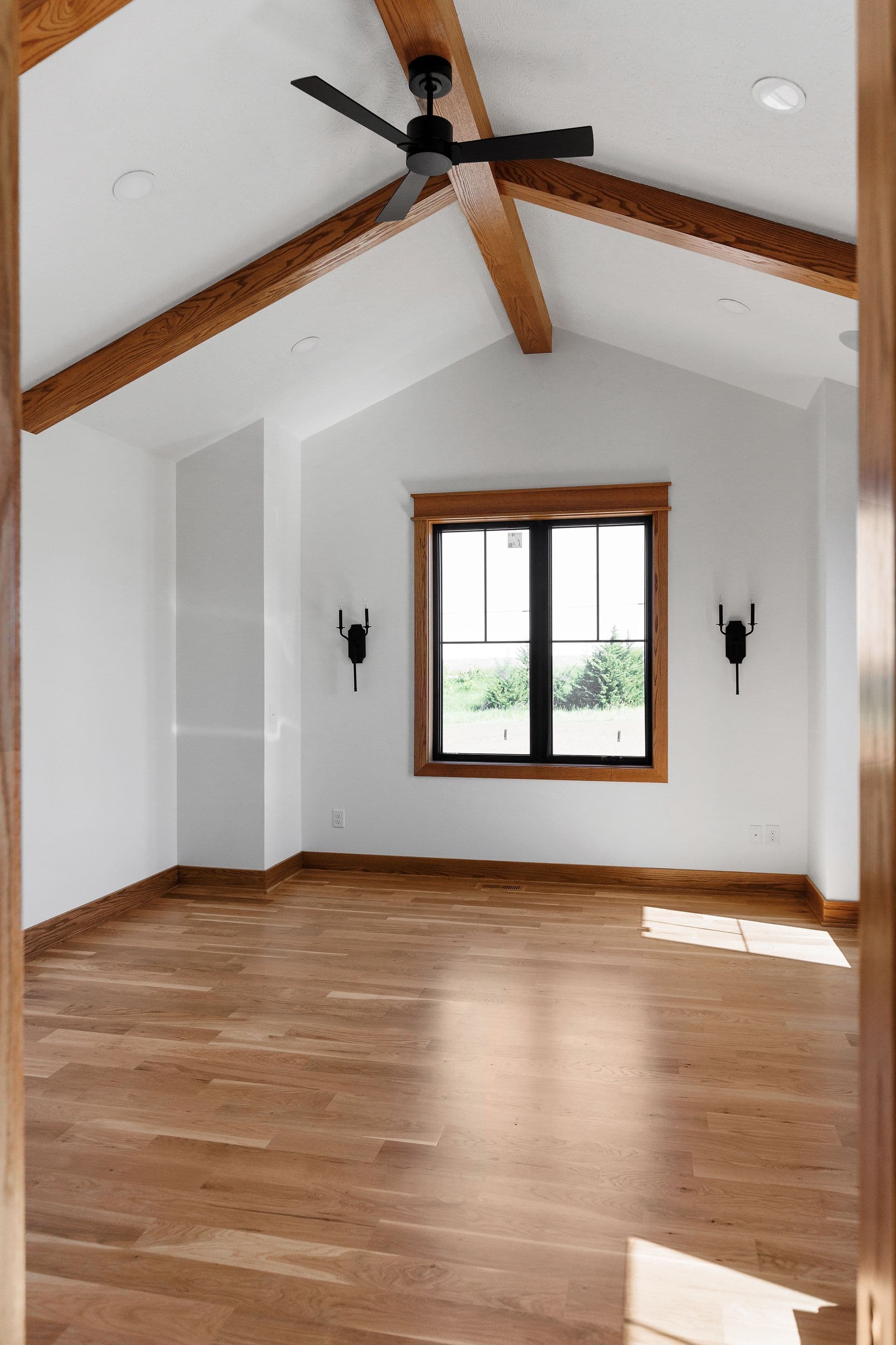 Modern bedroom featuring a vaulted ceiling with exposed wood beams, a black ceiling fan, and a window with black mullions. The space includes light hardwood flooring and dark sconces flanking the window.