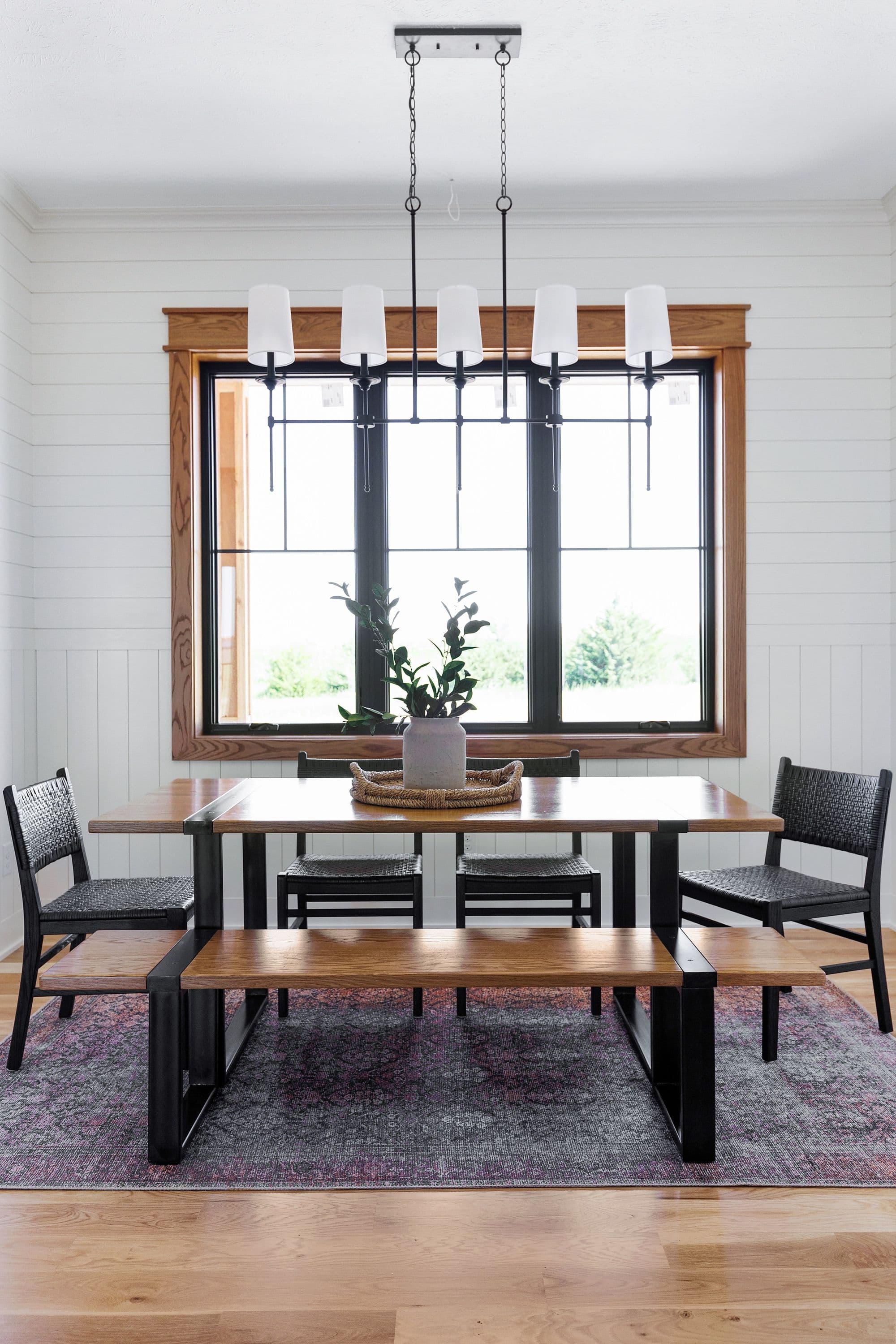Dining room features a wood table with metal legs, surrounded by woven chairs and a wooden bench. A modern chandelier hangs above the table, and a large window provides natural light.