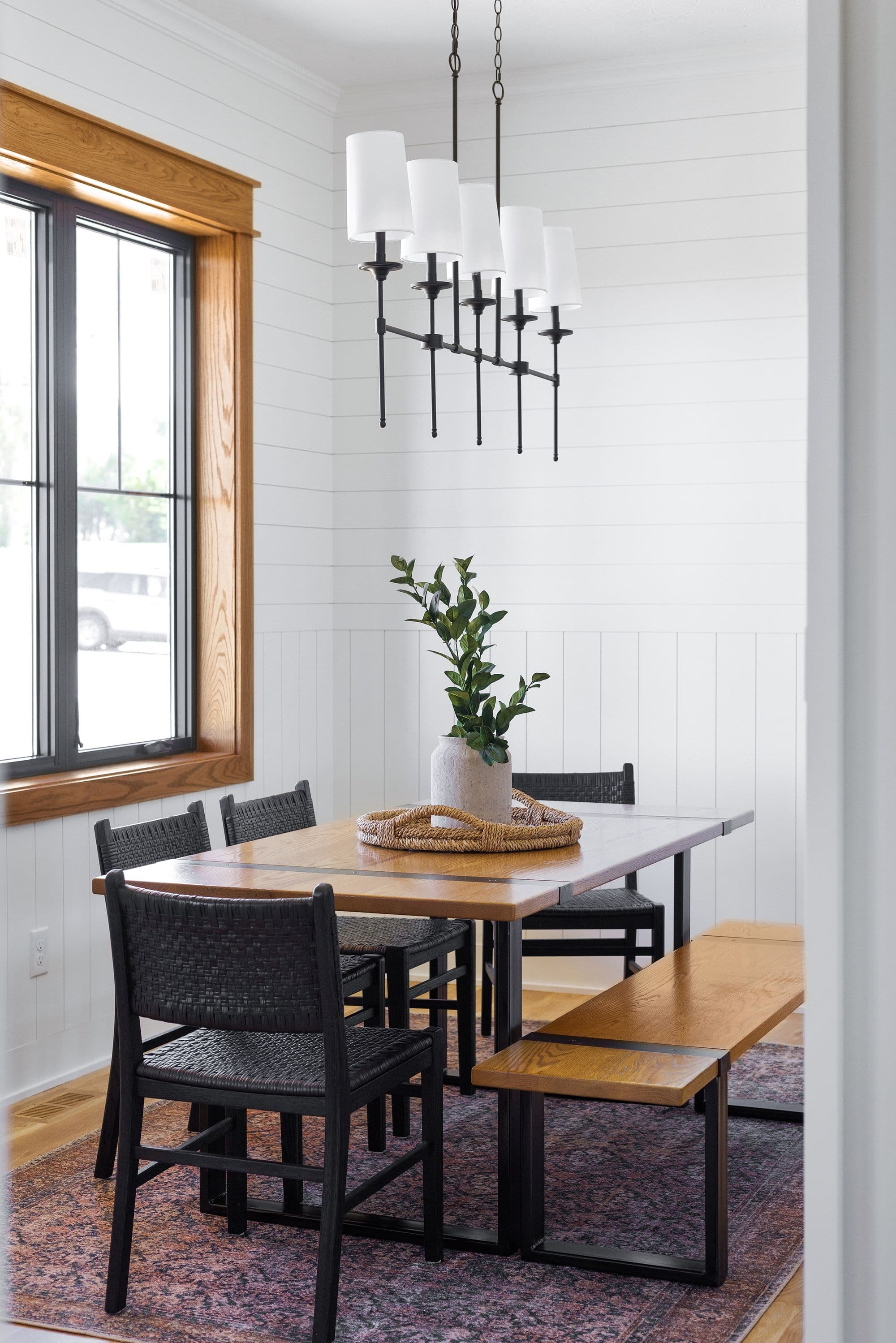 Dining room showcases a wooden table with black woven chairs and bench, complemented by a modern chandelier. The room features white shiplap walls and a window framed in natural wood.