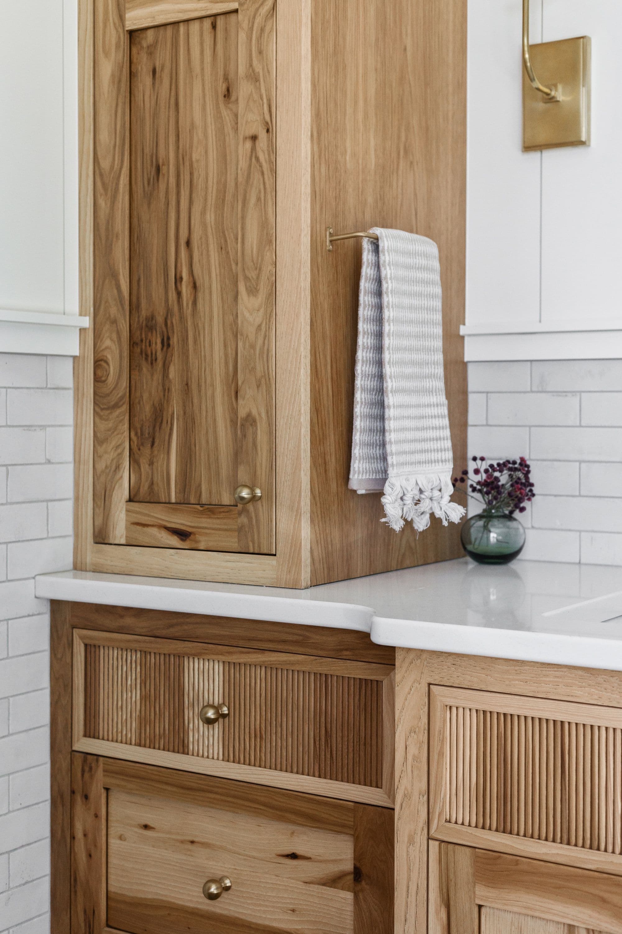Close up of a modern kitchen cabinet with white countertops and white subway tile backsplash, showcasing a natural wood finish and gold hardware. A light grey and white striped towel hangs next to a small vase.