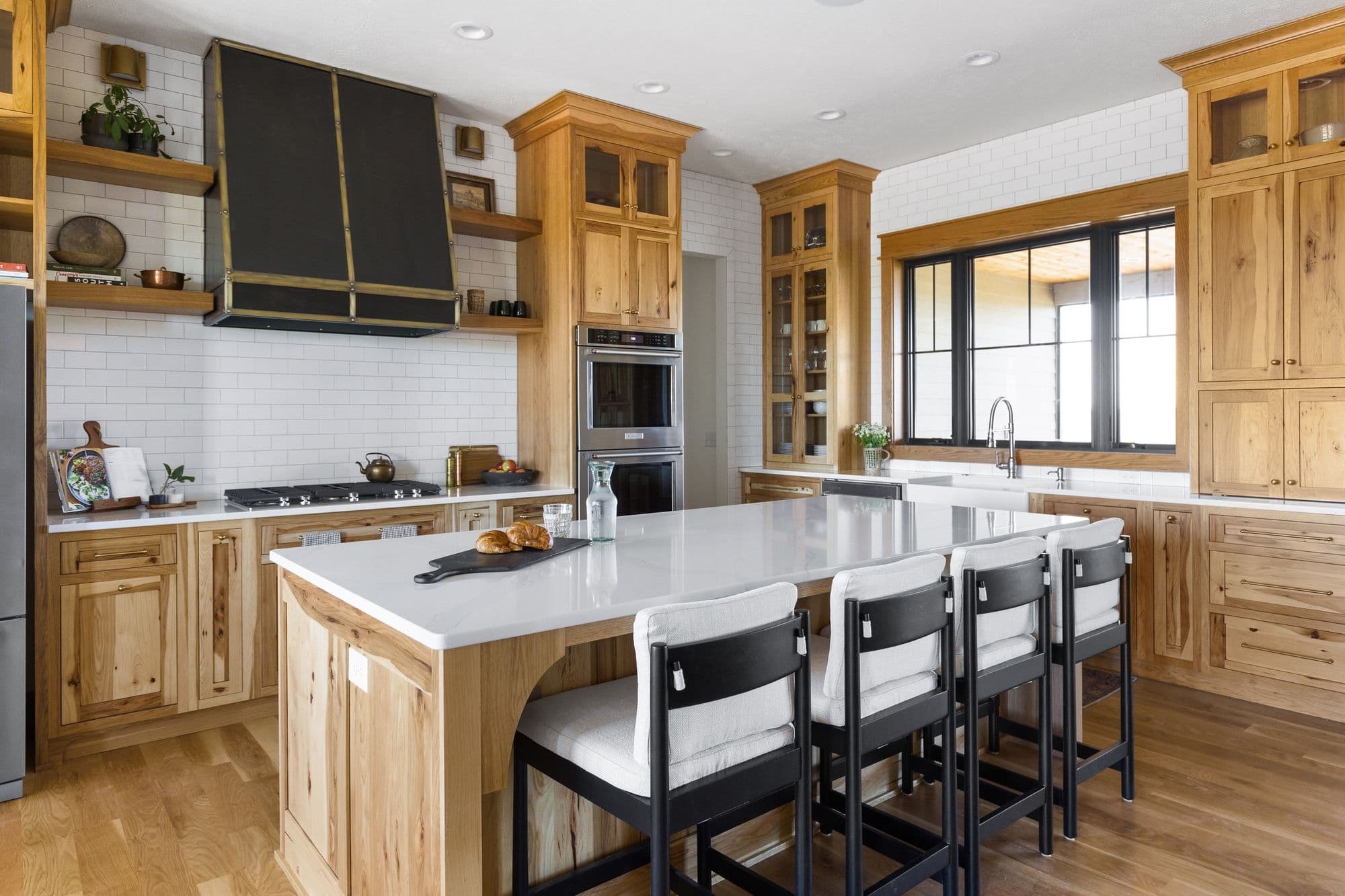 Bright kitchen showcasing light wood cabinetry, white countertops, and black accents in the vent hood and barstools. Subway tile backsplash and wood flooring complement the design.