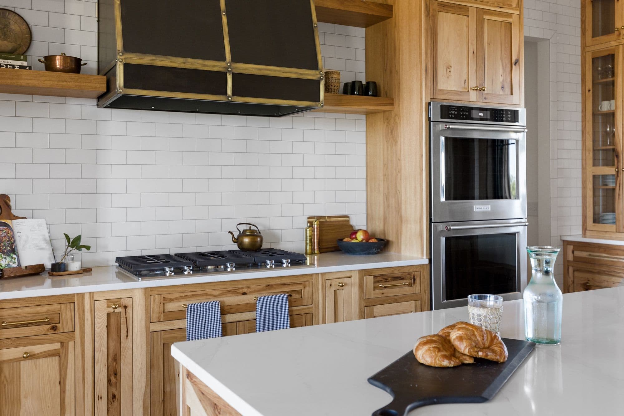 Warm-toned kitchen with white subway tile backsplash, wooden cabinets, and stainless steel appliances. A black cutting board with croissants sits on a white countertop.