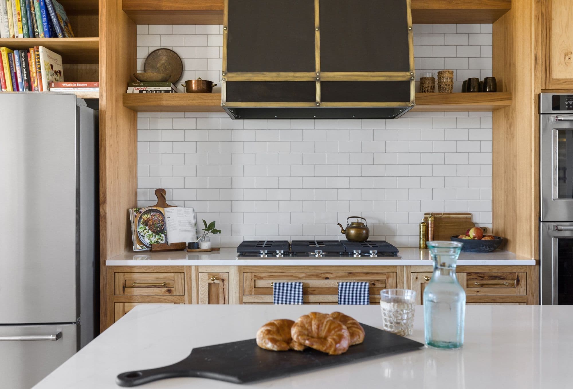 Well-lit kitchen with natural wood cabinetry and white subway tile backsplash. An island with a white countertop sits in the foreground, displaying a black cutting board with pastries, a glass of water and a blue water bottle.