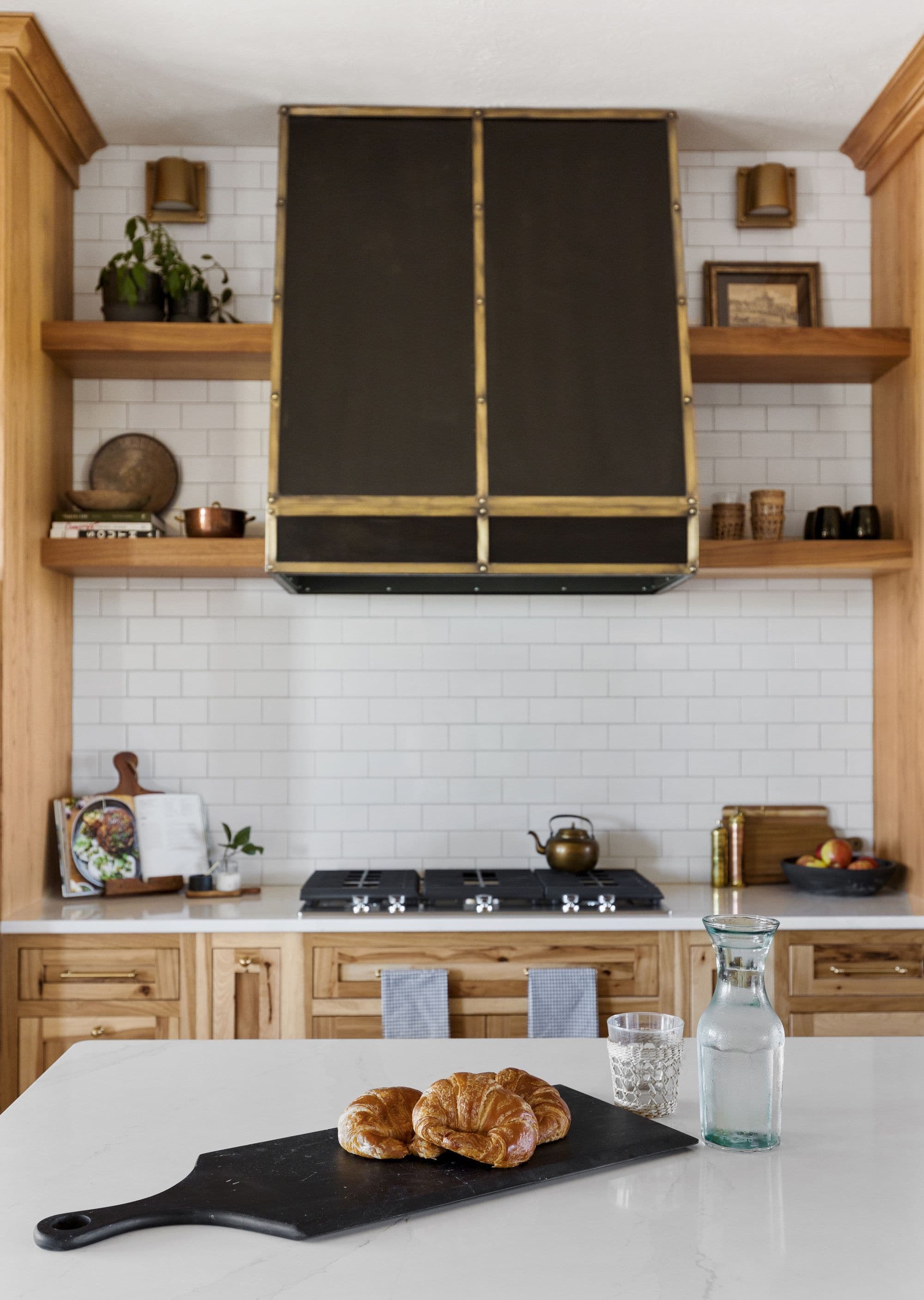 Modern kitchen featuring light wood cabinetry, a white subway tile backsplash, and a dark metal range hood with brass accents. A white countertop island displays croissants on a black cutting board, and a water carafe.