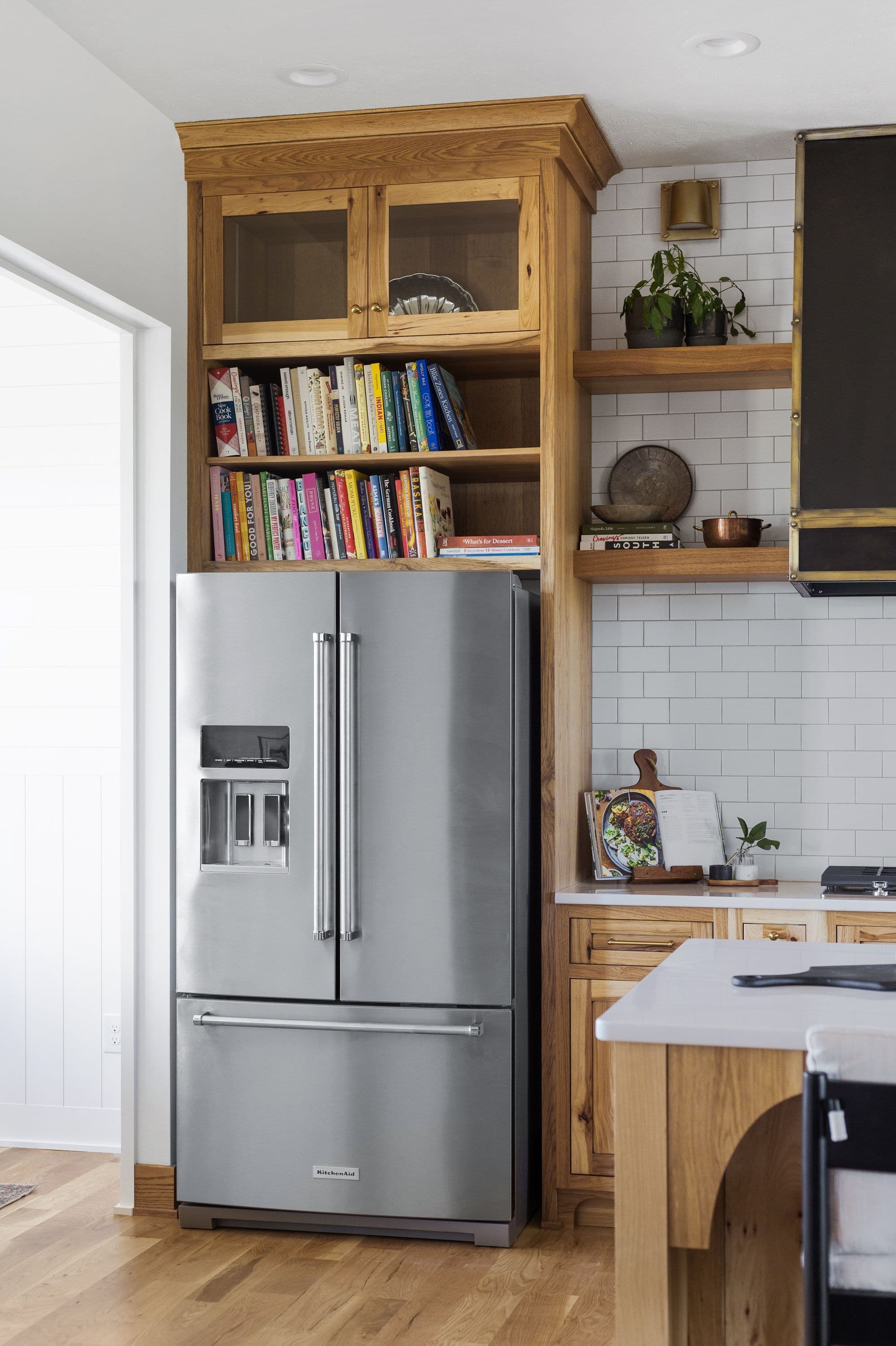 A well-lit kitchen features custom wood cabinetry around a stainless steel refrigerator. Open shelving displays cookbooks and decorative items, complementing the white subway tile backsplash.