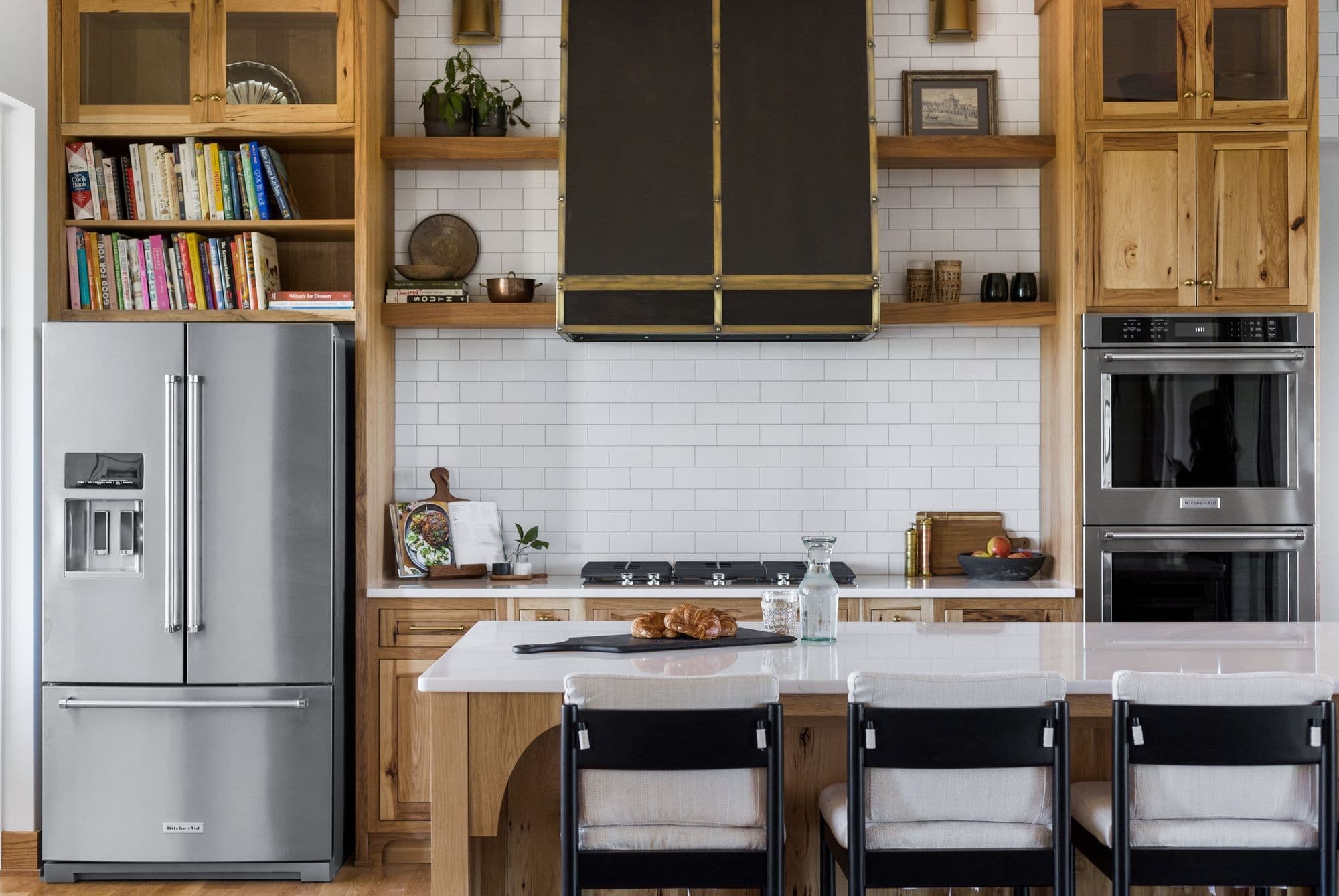 Modern kitchen features a stainless steel KitchenAid refrigerator and double oven, contrasted with warm wood cabinetry and open shelving displaying cookbooks and decor. A white quartz countertop island with black and white bar stools anchors the space, complemented by a white subway tile backsplash.