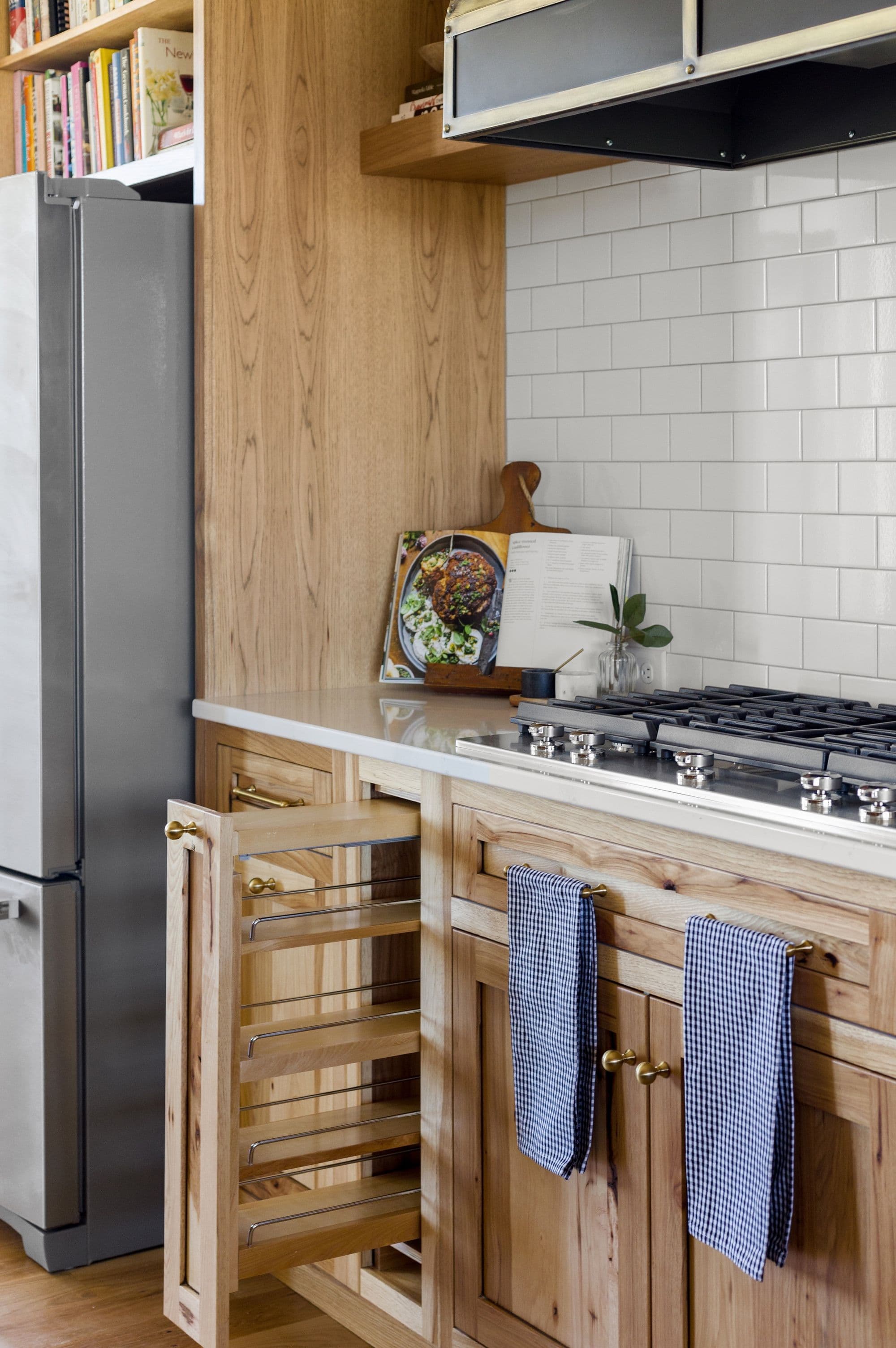 A modern kitchen features light wood cabinetry with gold hardware, a white countertop, and a gas range. Subway tile backsplash and a stainless steel refrigerator are also visible.