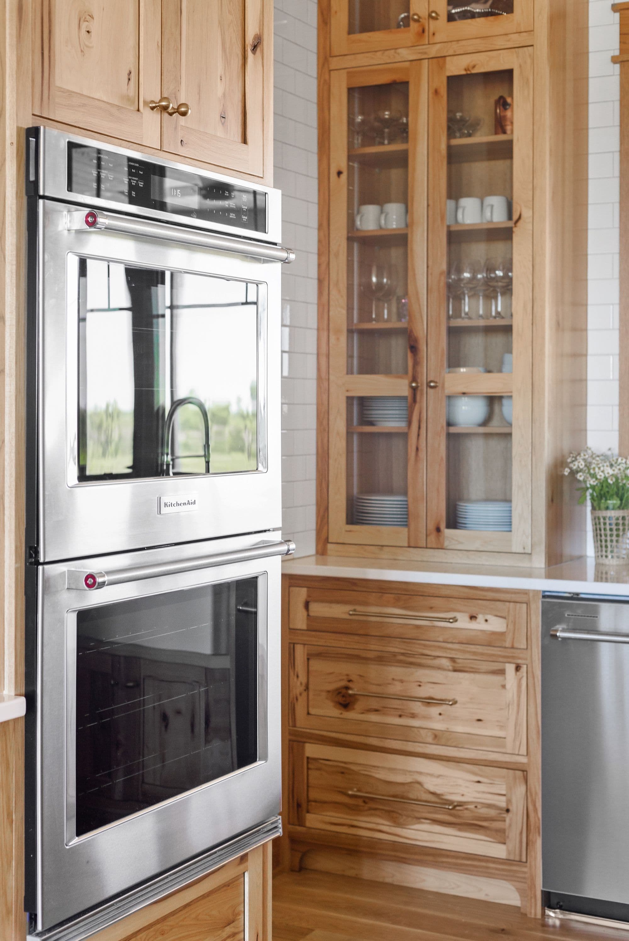 Kitchen area features stainless steel KitchenAid double ovens built into natural wood cabinets with brass hardware and glass-front upper cabinets showcasing dishware. White subway tile backsplash is visible above the white countertops.