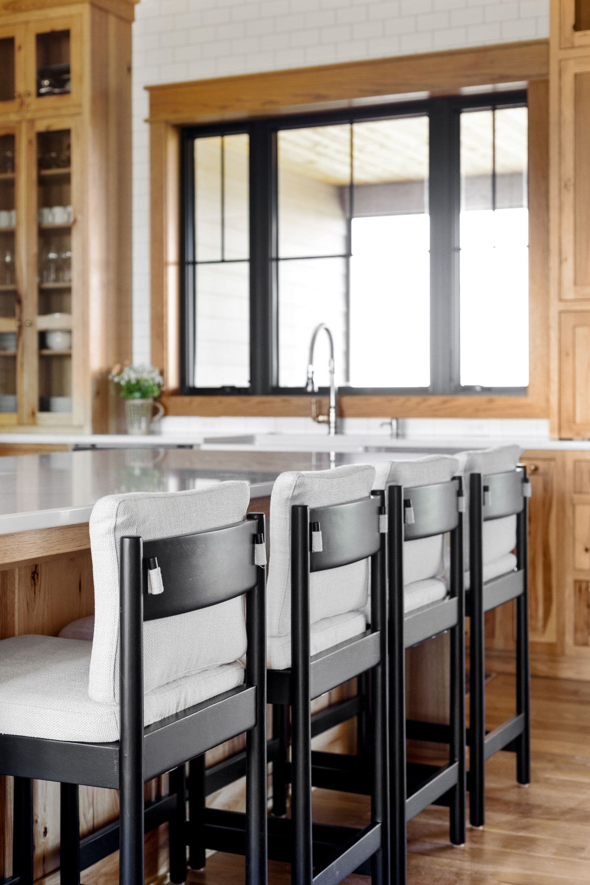 Modern kitchen island with four bar stools and a large window providing natural light. The kitchen features custom wood cabinetry and a white countertop.