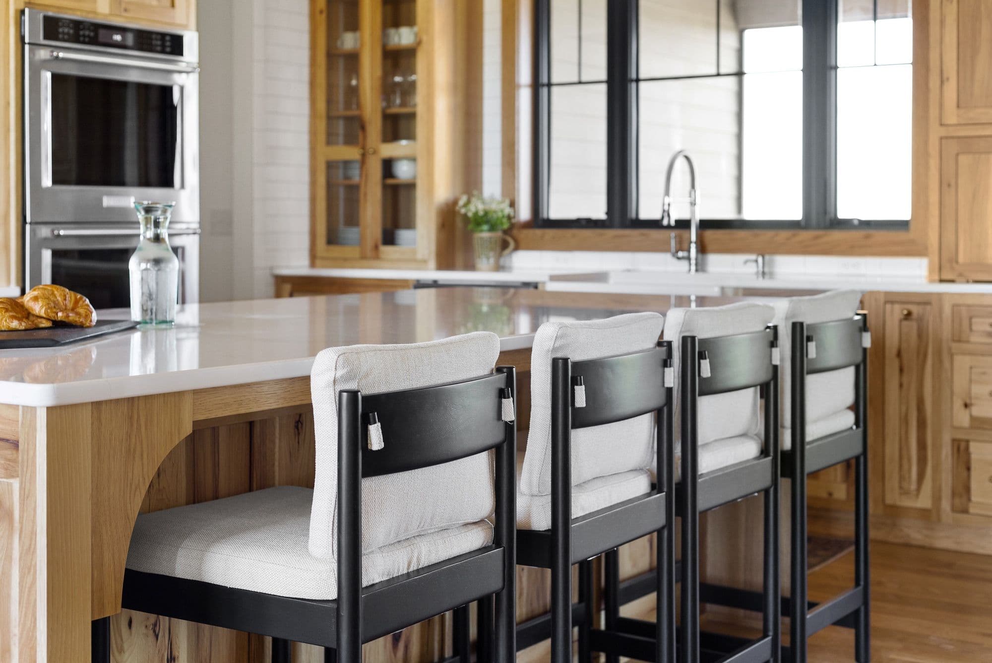 Bright kitchen with light wood cabinetry and a white countertop island features modern black barstools with white cushions. Stainless steel appliances are visible, and a large window provides natural light.