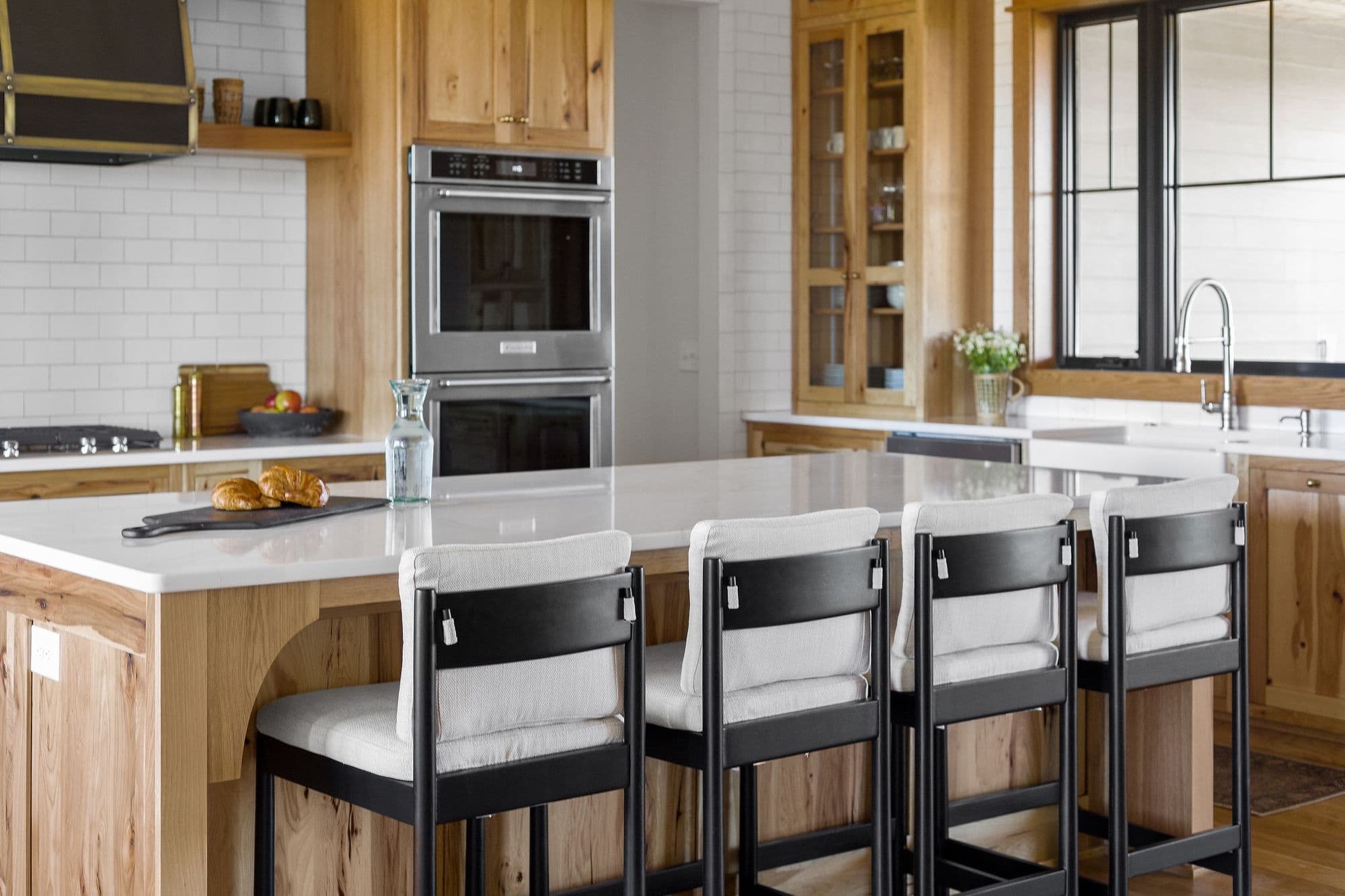 Bright kitchen featuring warm wood cabinetry, a white countertop island with four black and light-colored bar stools, and stainless steel appliances. The subway tile backsplash adds a clean, modern touch to the rustic design.