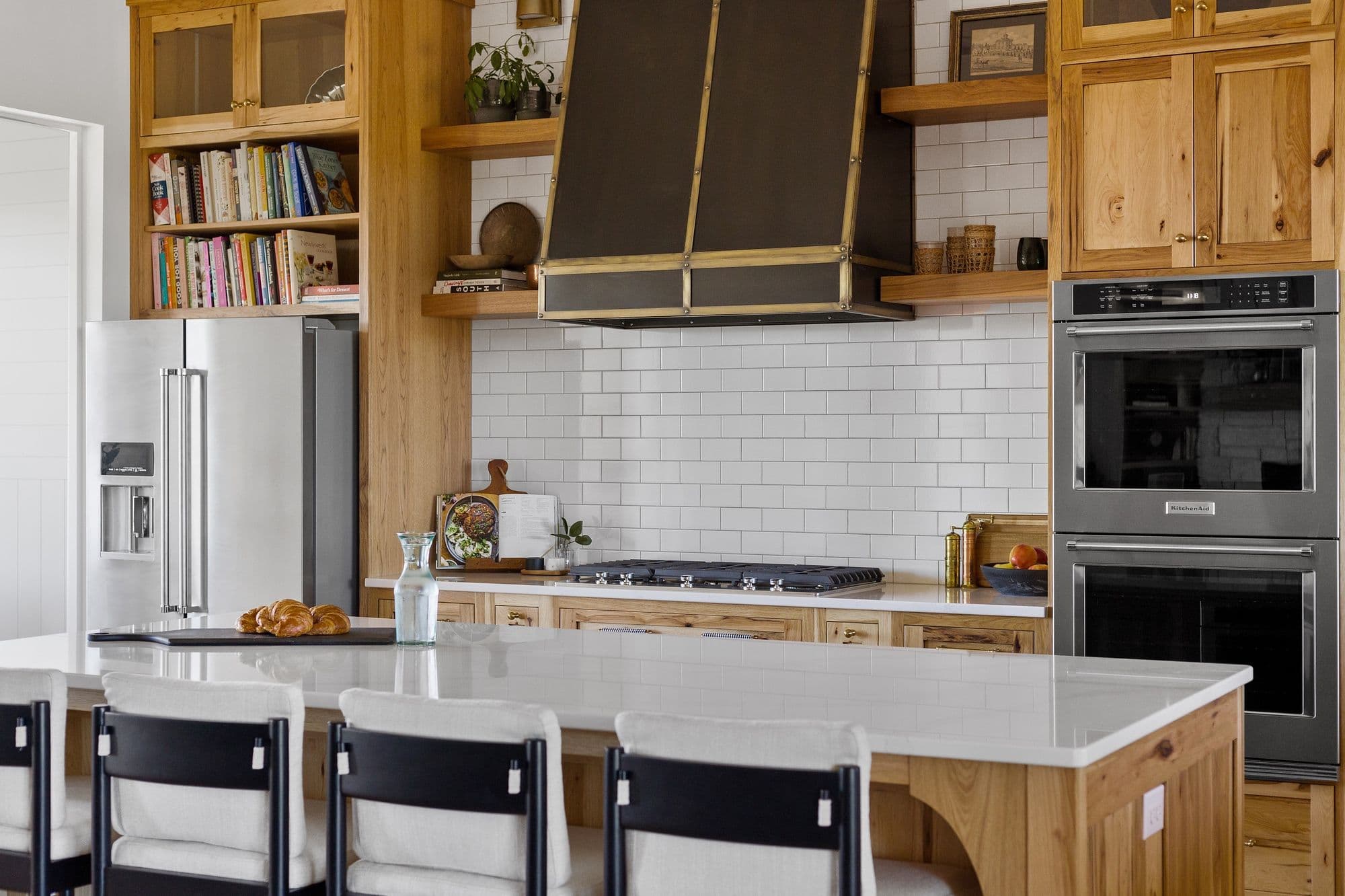 Well-lit kitchen featuring natural wood cabinetry, white subway tile backsplash, and stainless steel appliances. An island with white countertop and black chairs sits in the foreground, with croissants and water carafe.