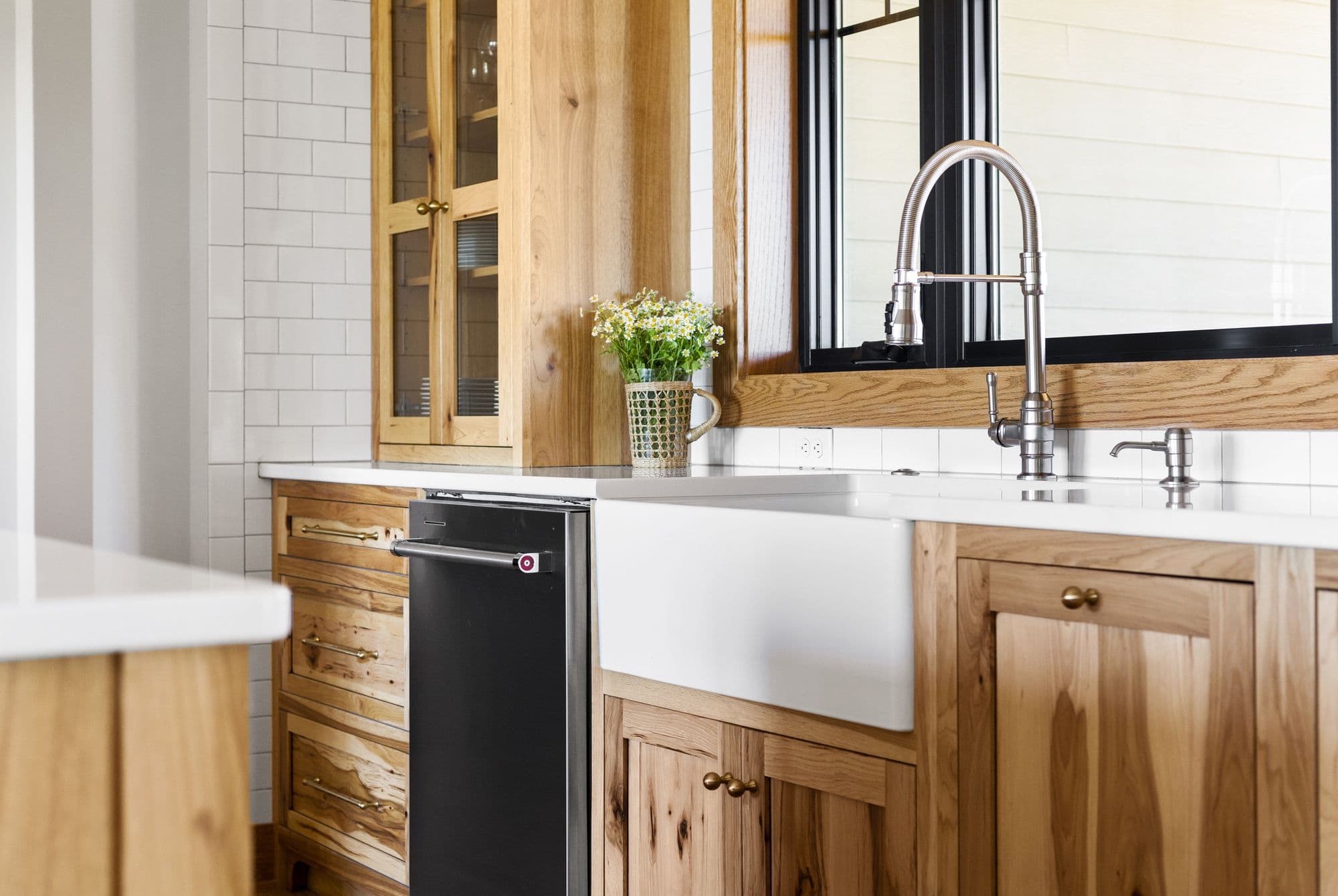Modern kitchen features light wood cabinetry with brass hardware, a white farmhouse sink, and a black dishwasher. Subway tile backsplash complements the light countertops and a stainless steel faucet adds a sleek touch.