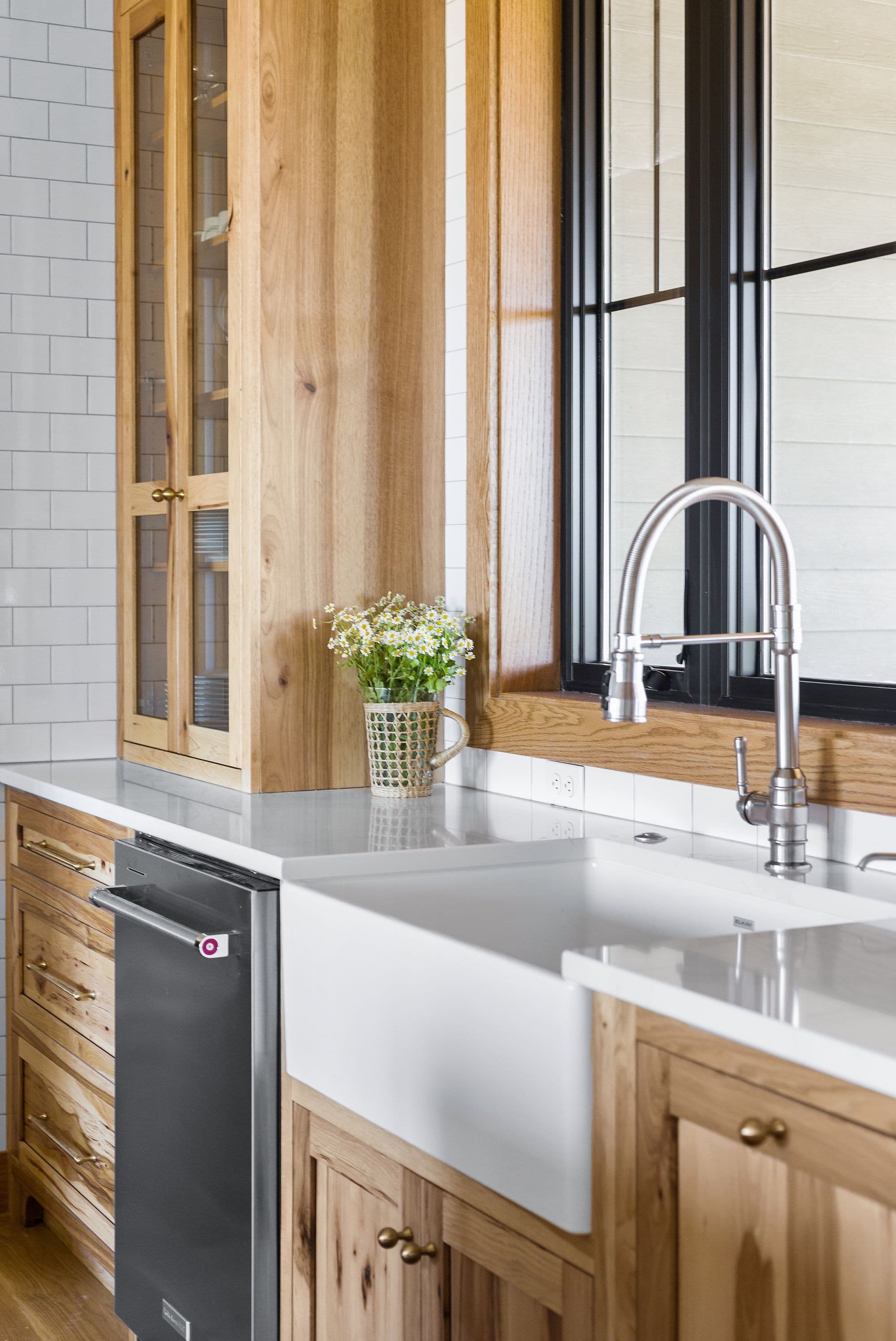 Modern kitchen features natural wood cabinetry with brass hardware, white farmhouse sink, and black framed window. Subway tile backsplash and light gray countertops complement the design.