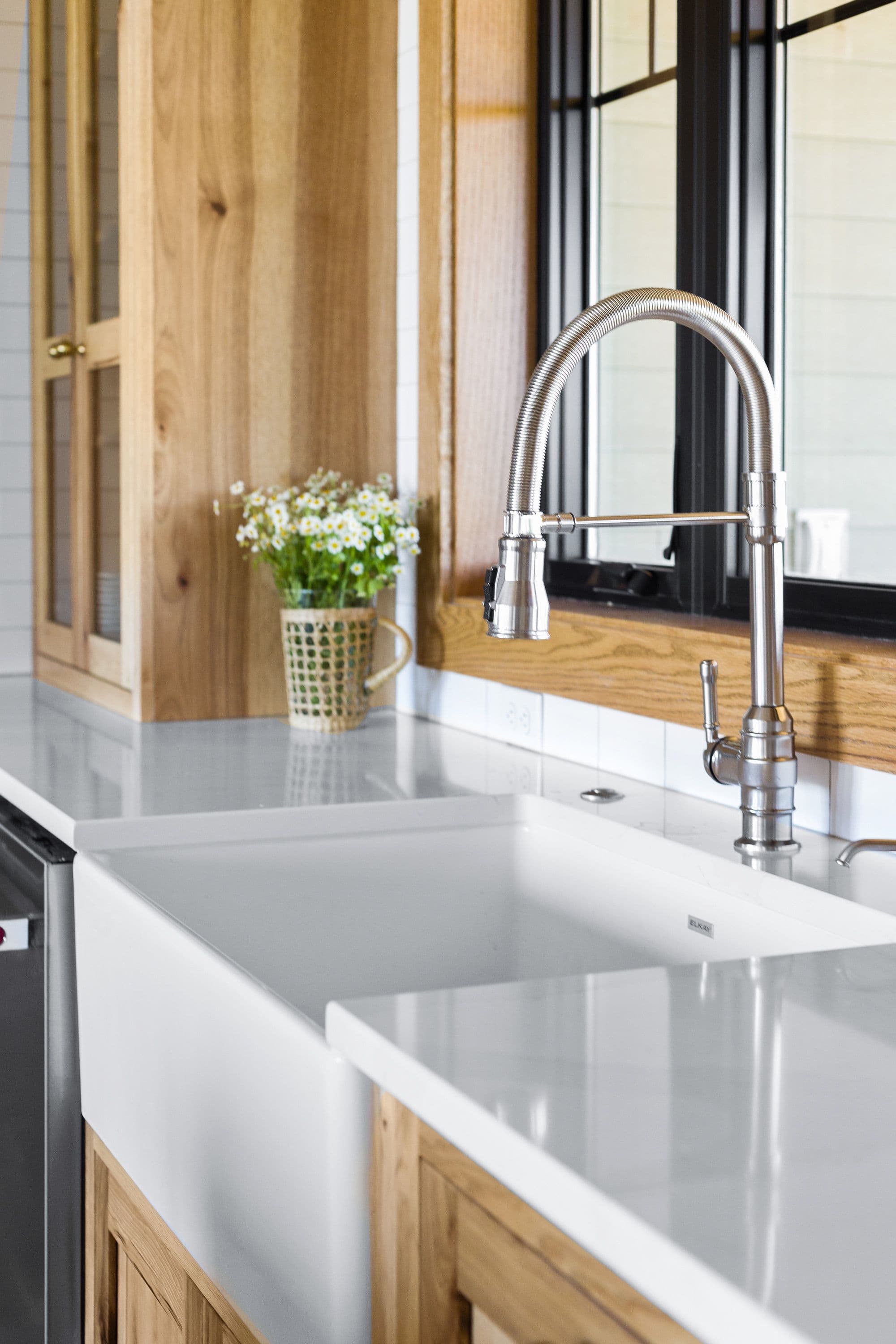 Farmhouse-style kitchen featuring a white apron-front sink with a stainless steel faucet, white subway tile backsplash, and light wood cabinets. Fresh daisies in a wicker basket add a touch of nature to the countertop.