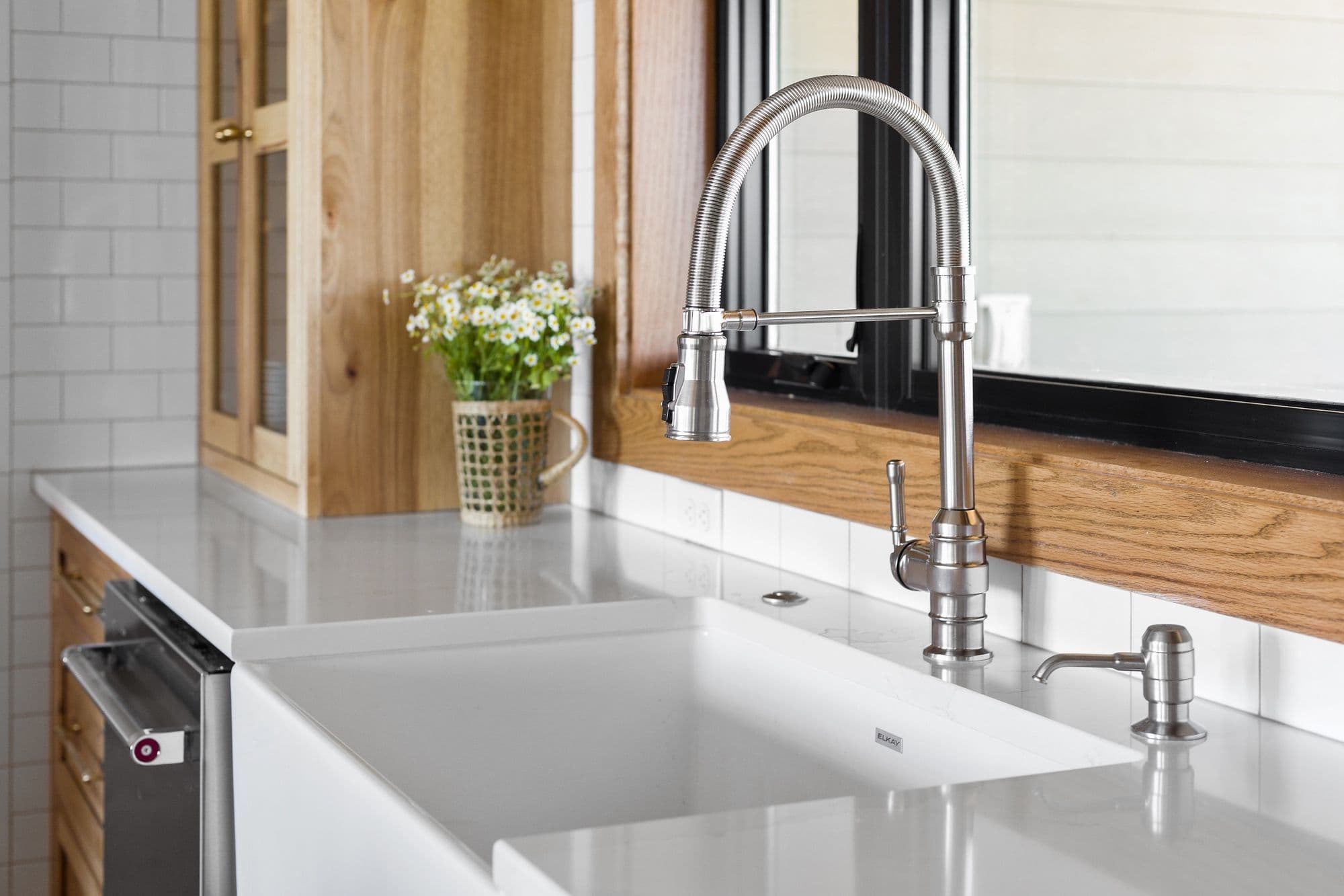 Modern kitchen features a white farmhouse sink, stainless steel faucet, and light gray quartz countertops. A window with a dark frame is centered above the sink, adding natural light to the space.