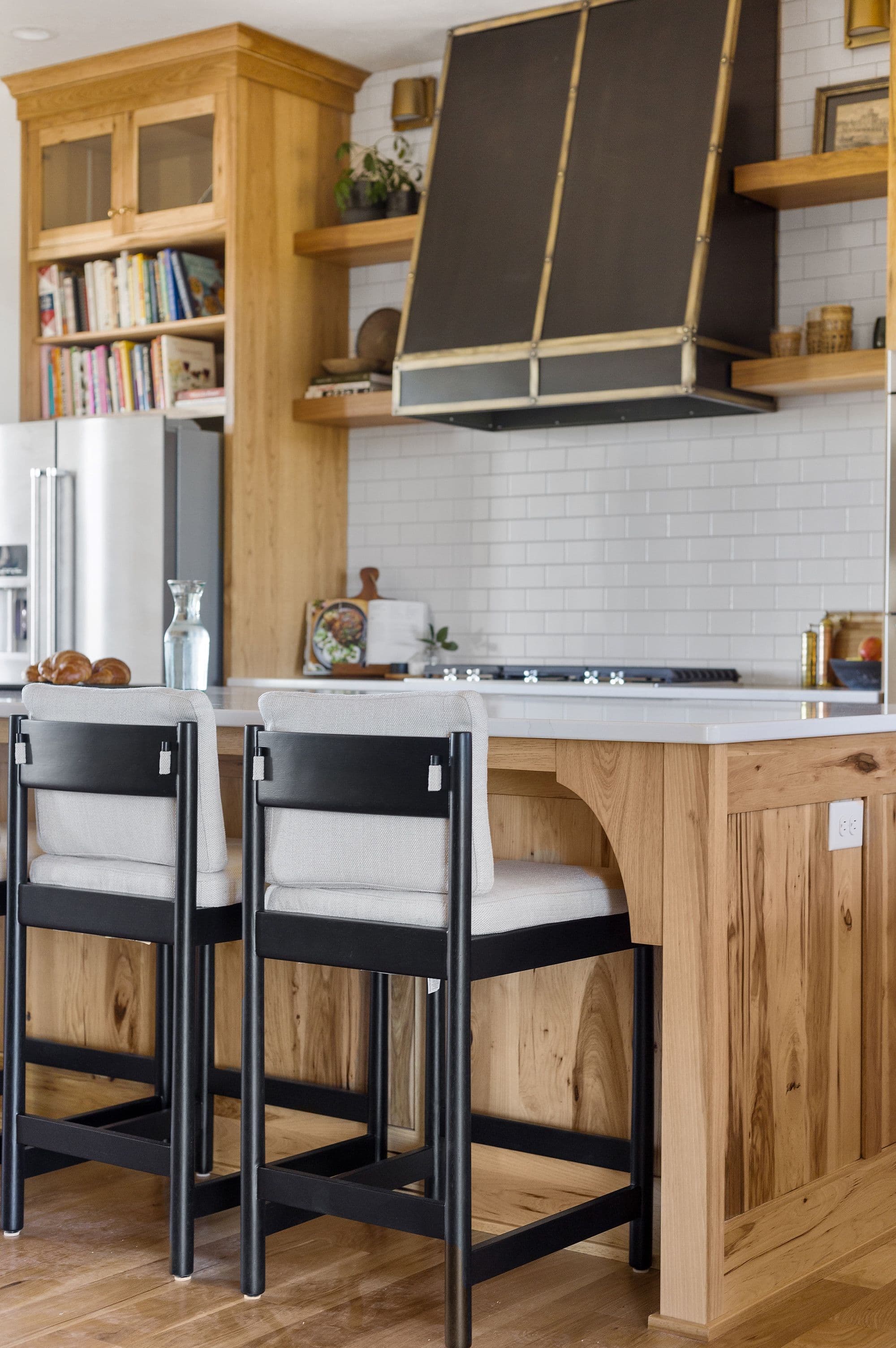 Modern kitchen with light wood cabinetry, black metal range hood, and white subway tile. Two counter stools with light upholstery and black frames sit at the kitchen island.