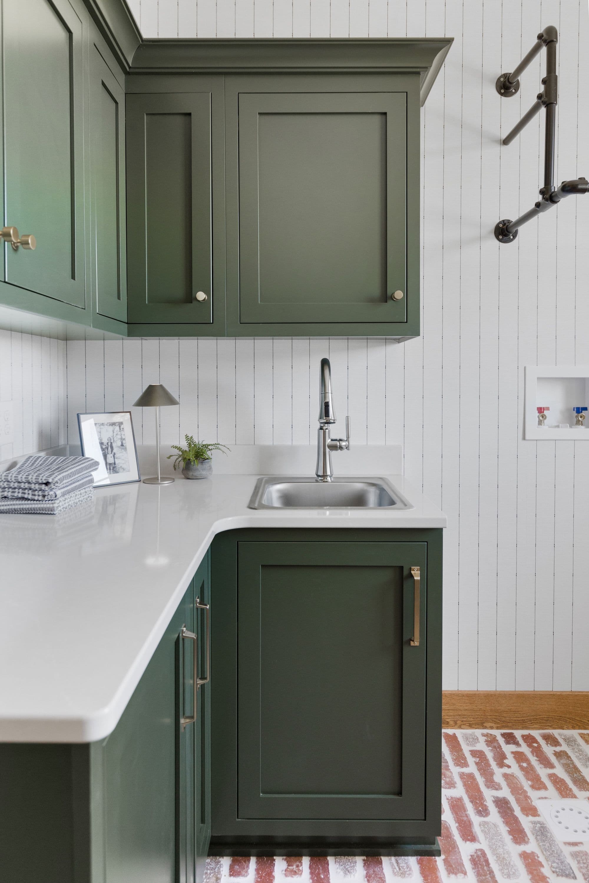 Laundry room features dark green cabinets, a stainless steel sink, and a white countertop. The floor is red brick and the walls are adorned with wallpaper, complemented by a black pipe drying rack.