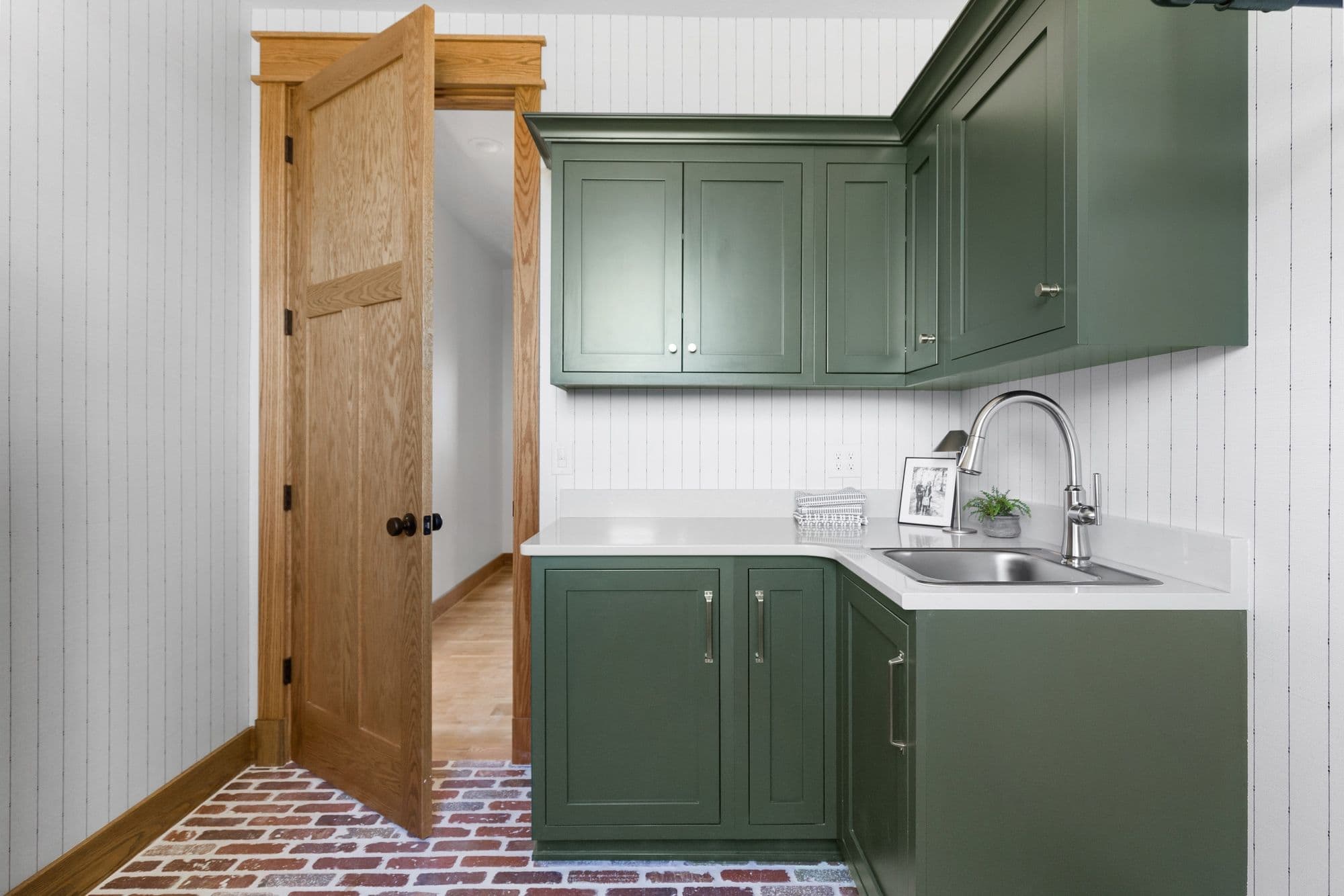 Laundry room with brick flooring and olive green cabinets. A wood door leads into an adjacent room with light wood flooring.